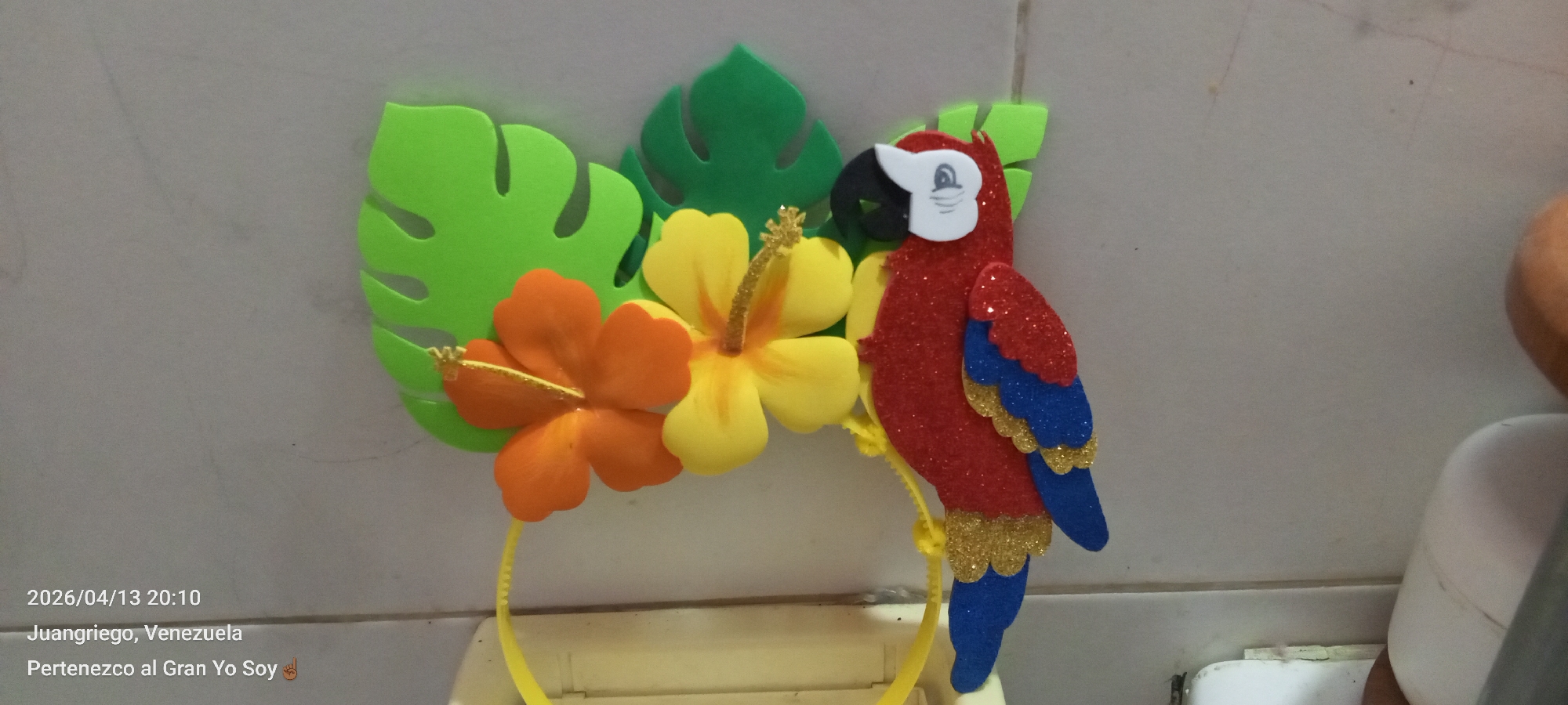 A vibrant, handcrafted tropical-themed headband is the primary subject of the image. It features green foam Monstera leaves, an orange foam hibiscus flower, a yellow foam hibiscus flower, and a red foam parrot with blue and gold accents. The headband is yellow.
The photograph was taken in Juangriego, Venezuela, according to the timestamp, but not specifically in Altagracia.
The background appears to be a plain, light-colored surface, likely a wall or countertop. The lighting is somewhat diffuse.