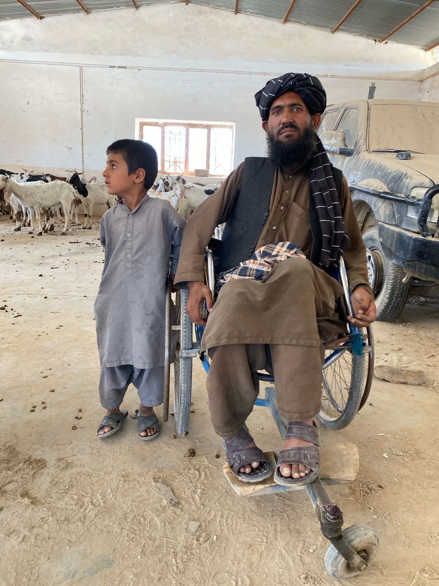 The image shows a man in a wheelchair and a young boy standing next to him, in what appears to be a barn or shed filled with goats. The man is wearing a dark turban, a dark vest over a brown shirt and pants, and sandals. He has a beard. The boy is wearing a grey tunic and pants, and sandals. In the background, there are numerous white and black goats. A portion of a dusty, light-colored vehicle is visible to the right of the man. The floor is dirt and scattered with debris. The location is identified as the city of Pasab, Afghanistan.