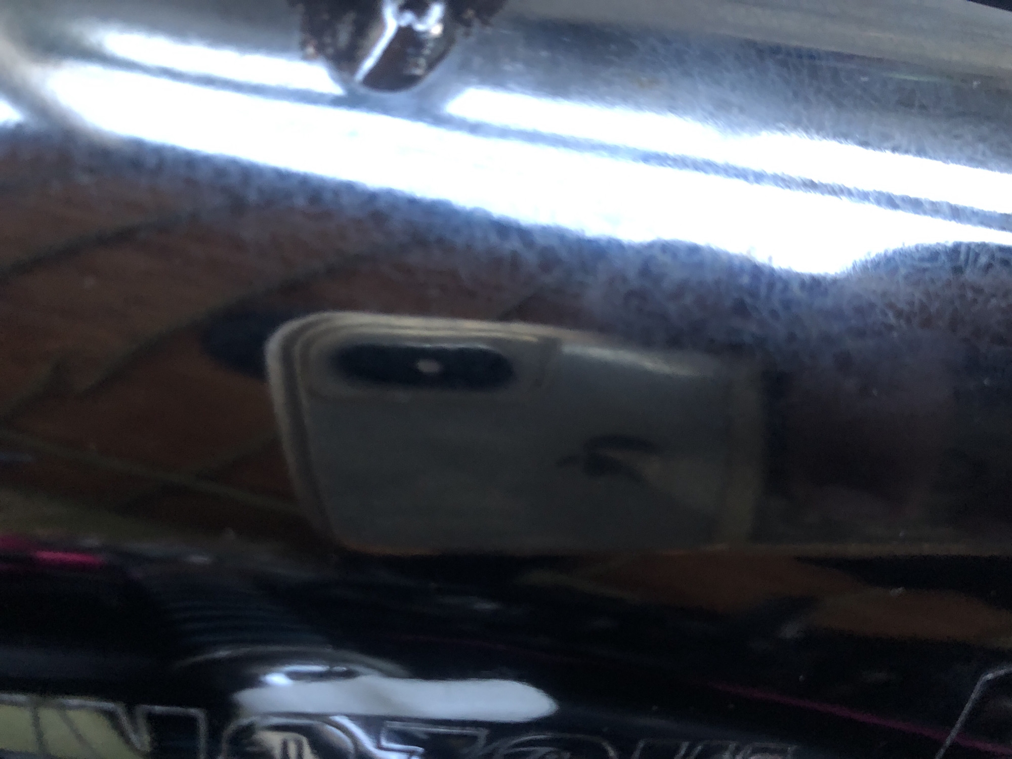 The image shows a reflection of a cell phone on a shiny surface, likely chrome or polished metal. The cell phone has a light-colored case and is reflecting back the camera lens. The surrounding environment is blurry and indistinct. There is no information available to identify the location as Altagracia, Venezuela.