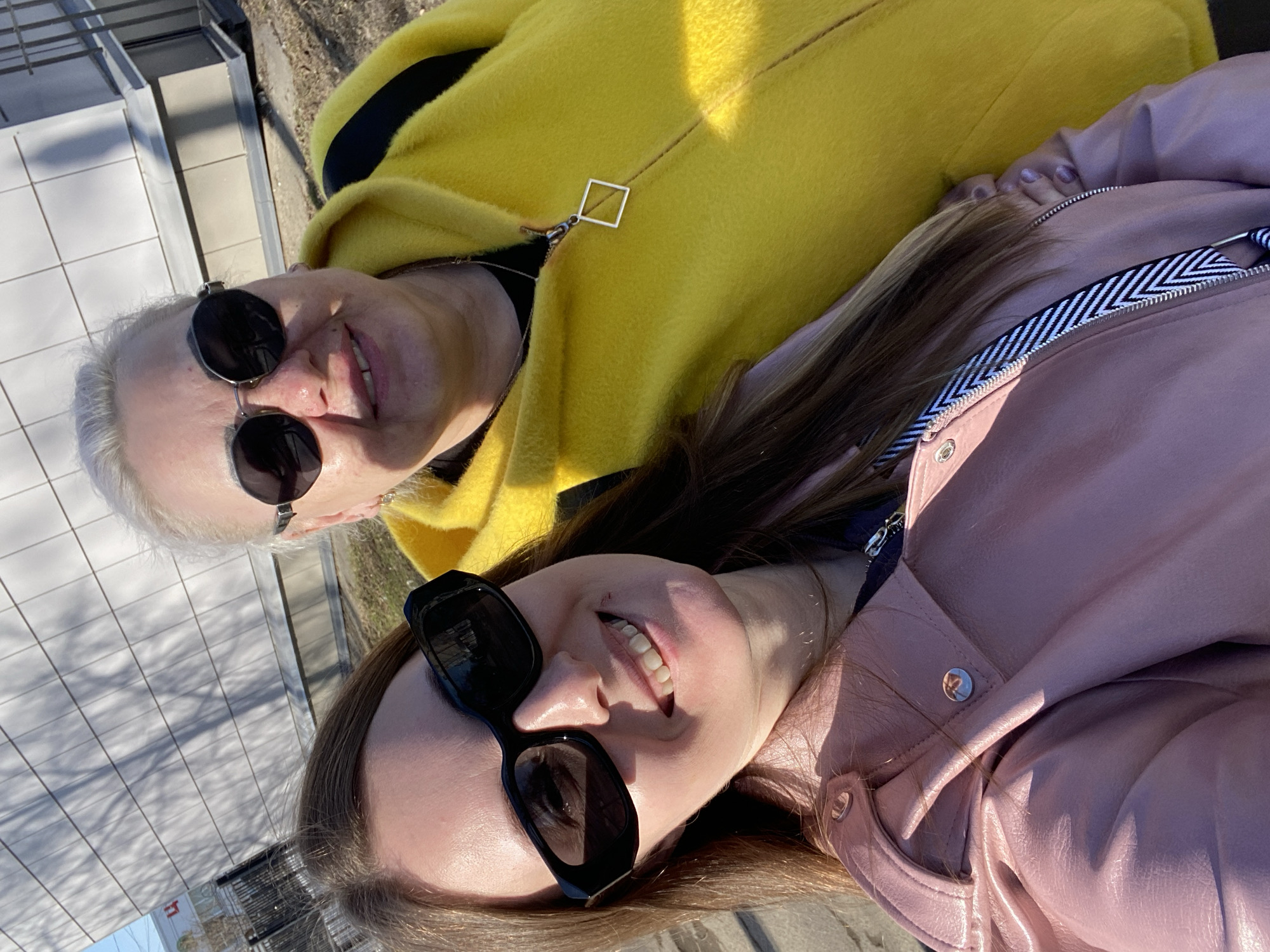 Two women are posing outdoors for a photo. The woman in the upper left wears a bright yellow jacket with a black stripe and silver square zipper pull. She has short white hair and dark sunglasses. The woman in the lower right has long brown hair and is wearing large dark sunglasses and a pink leather jacket with a black and white striped zipper. Both women are smiling and appear to be lying down, possibly on a sidewalk or paved area. The location appears to be Chaikovsky, Russia. A building and trees can be seen in the background.