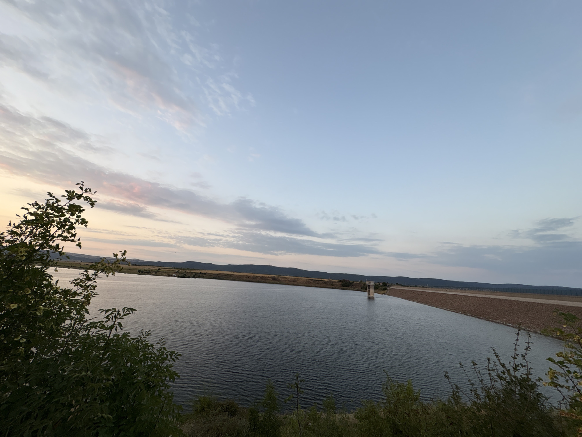 The image depicts a wide expanse of water, likely a reservoir or lake, at dusk. The water's surface is rippled, reflecting the soft light of the setting sun. In the distance, a long dam structure is visible, with a tall tower-like building standing in the water near the dam. The sky is a gradient of soft blues and pinks, with scattered clouds. The foreground features dark green foliage, suggesting trees or bushes. The surrounding landscape appears to be rolling hills covered in dry vegetation, hinting at a rural or semi-arid environment. The scene is serene and captures a natural landscape. This scene is likely the Tankovo Reservoir, located near the village of Tankovo in Bulgaria.
