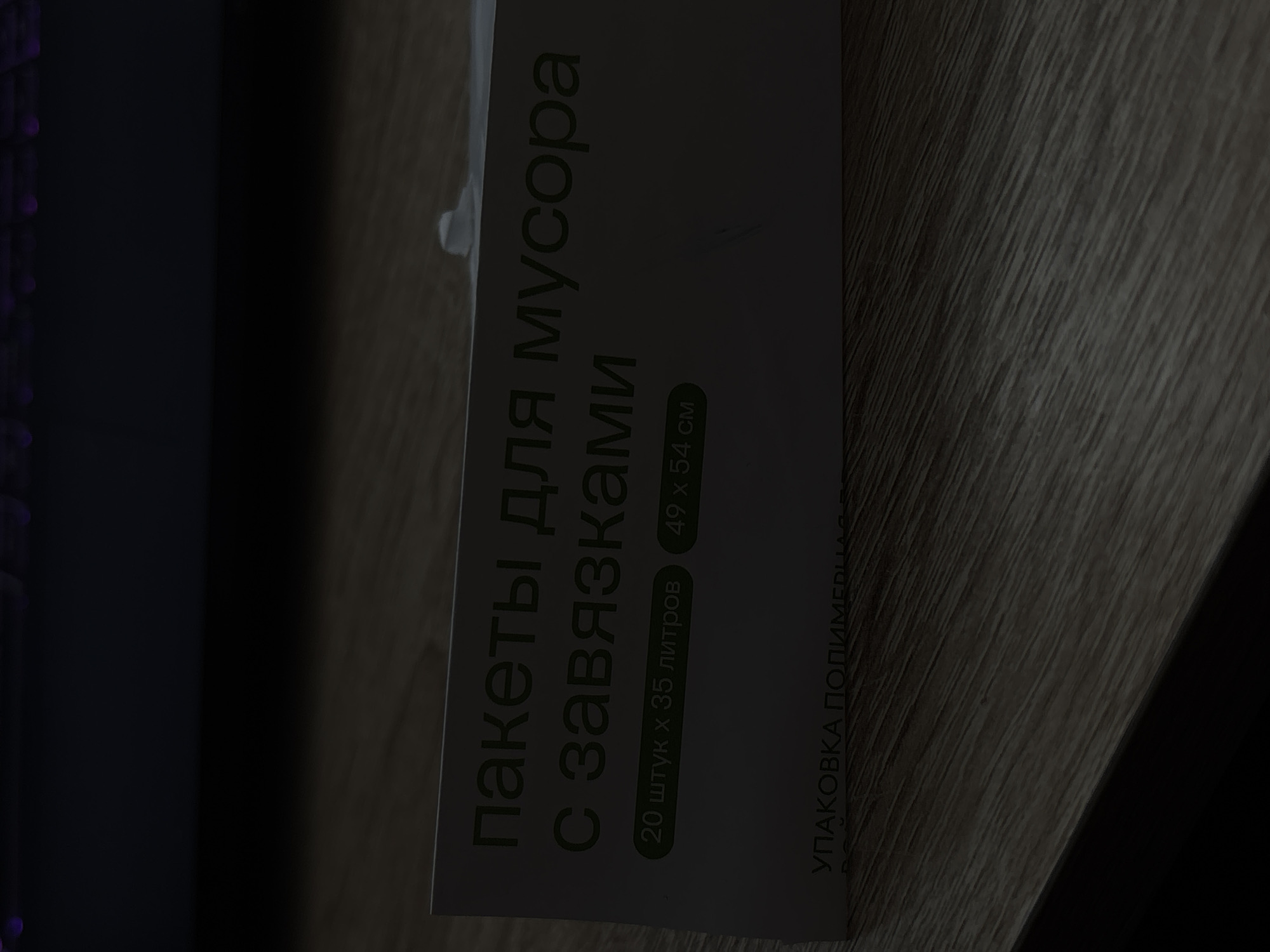The image shows a close-up of a white cardboard box with text in Russian. The text reads "ПАКЕТЫ ДЛЯ МУСОРА С ЗАВЯЗКАМИ" which translates to "TRASH BAGS WITH TIES". Below this, there are two circular labels: "20 штук х 35 литров" (20 pieces x 35 liters) and "49 х 54 см" (49 x 54 cm). At the bottom of the box, partially obscured, is the text "УПАКОВКА ПОЛИМЕРНАЯ" (POLYMER PACKAGING). The box is placed on a dark wooden surface, possibly a table. The lighting is dim, casting shadows. There is no indication of a specific location in Saint Petersburg, Russia, or any people present in the image.