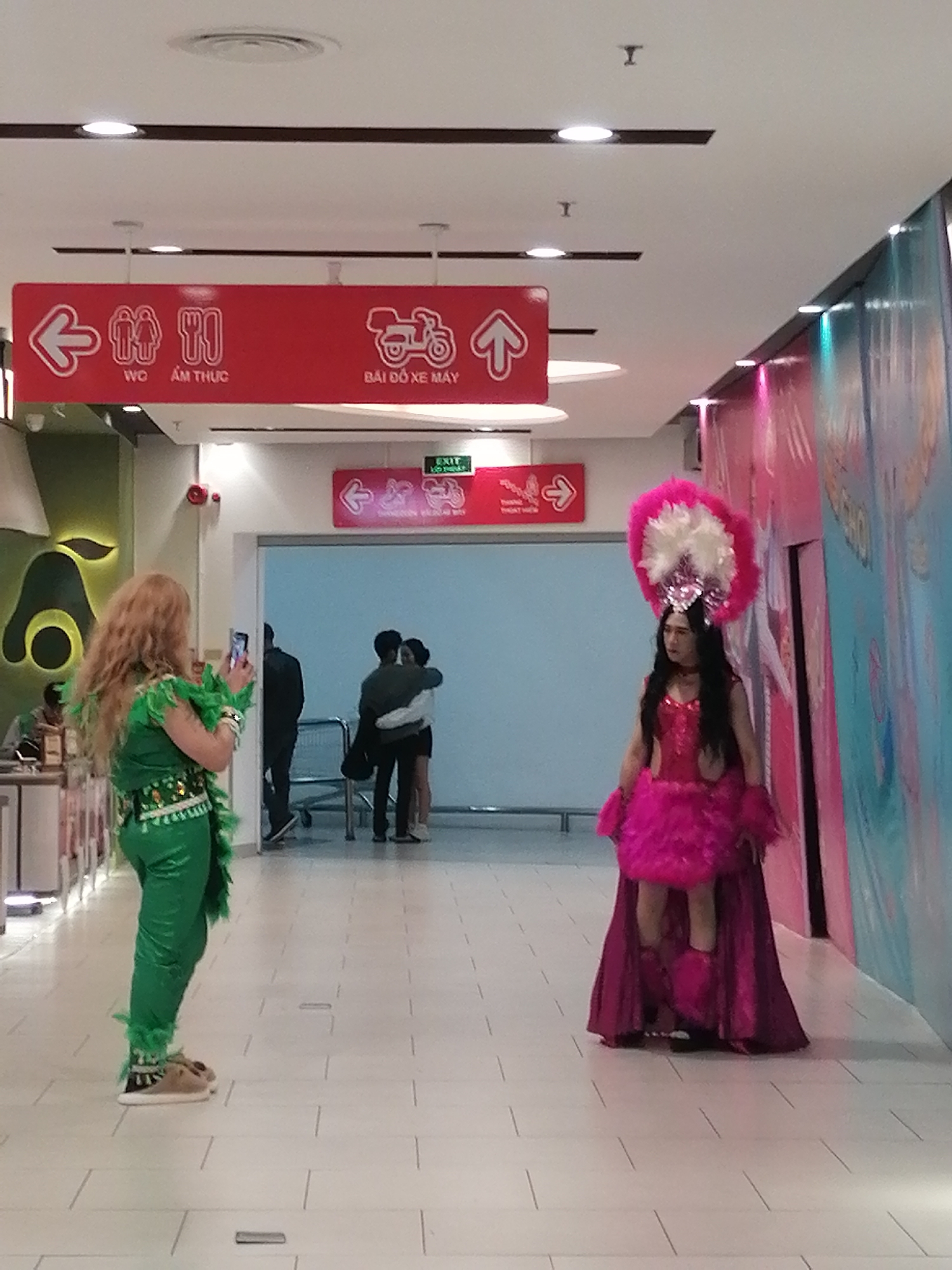 The image was captured in an indoor location, likely a shopping mall or commercial building, in Da Lat, Vietnam. Signage in Vietnamese indicates directions to restrooms (WC), food services (Ẩm Thực), and motorcycle parking (Bãi Đỗ Xe Máy).
Two individuals in extravagant costumes dominate the scene. On the left, a person wearing a green sequined outfit and a feathery boa appears to be taking a photo with a smartphone of another person in a pink sequined dress, feather headdress, and a long, flowing magenta cape. This second individual is posing for the photograph.
In the background, a couple can be seen embracing near an entrance or exit with signage reading "Exit." Two other individuals are walking through this entrance. The interior design includes modern lighting fixtures, tiled flooring, and a large decorative wall feature with vibrant colors.