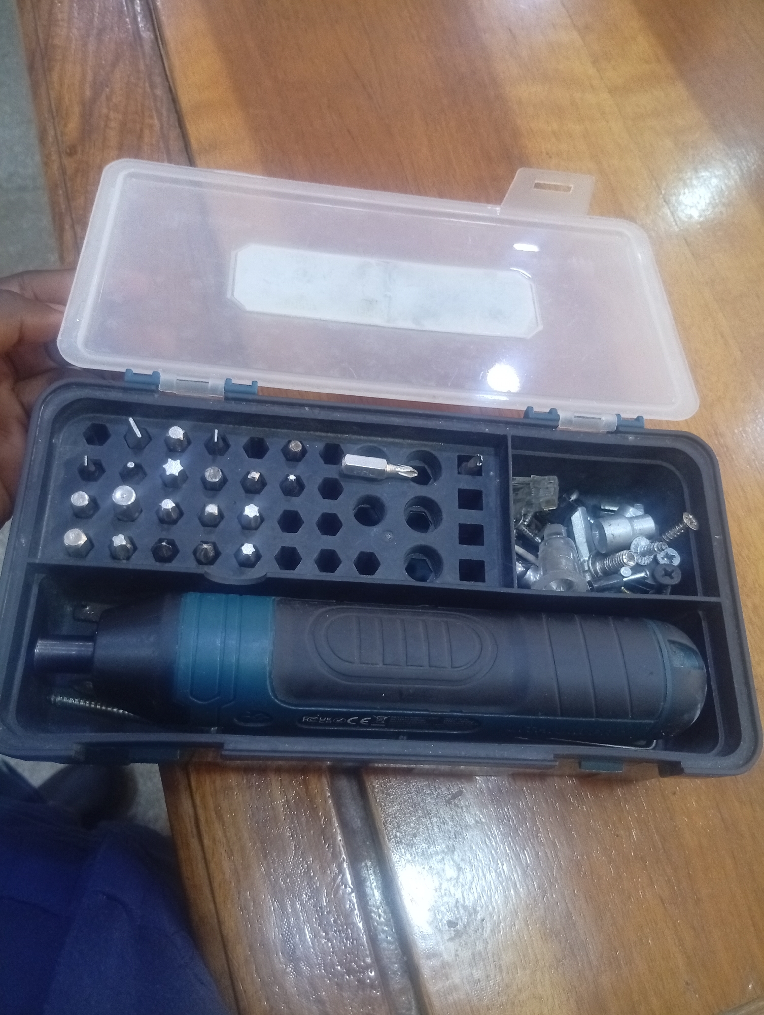 This media file shows an open toolbox containing a small electric screwdriver and a variety of screwdriver bits. The toolbox is made of black plastic with a clear plastic lid. The screwdriver has a black and teal handle. The bits are organized in a grid pattern within the toolbox. There is also a separate compartment in the toolbox containing various screws and other small metal parts. The toolbox is resting on a wooden surface, possibly a table. The scene appears to be indoors, given the lighting and the reflective quality of the wooden surface, which might be polished or wet. There are no people visibly active in the image, but a hand is partially visible at the top left, suggesting someone is holding or interacting with the toolbox. The overall impression is one of a tool kit ready for use or just being organized. Visible text on the screwdriver includes "FC UK CA CE". The location context provided is Shiroro, Nigeria, which suggests this is a view of tools possibly used for repair or maintenance work in that region. The time of day and weather conditions are not discernible from the image.