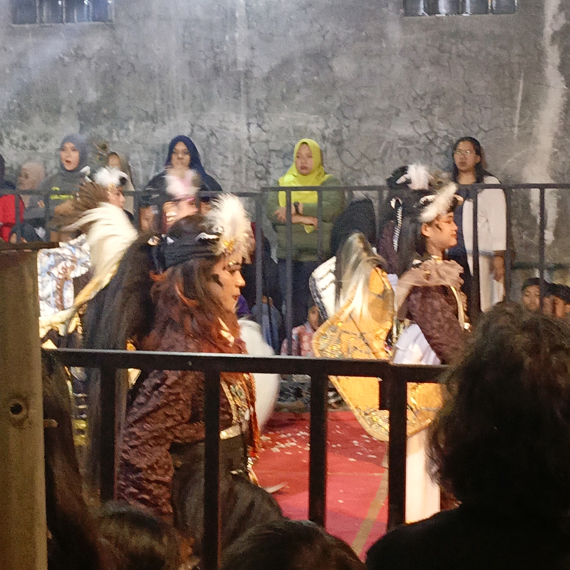 The image captures a lively scene, possibly a performance or cultural event, taking place outdoors in Salatiga, Indonesia. The backdrop is a textured, grey wall, suggestive of a natural stone or rough concrete surface. In the foreground, a black metal railing divides the scene, creating a sense of separation between the viewer and the participants.
Several individuals are visible, some in costume. On the left, a person in a dark, patterned outfit with elaborate headwear, featuring white feathers, is partially obscured by the railing. To their right, another individual wears a similar feathered headdress and a layered costume with metallic gold and orange accents. Further back, several women are seen, some wearing hijabs in various colors like blue, grey, and bright yellow. One woman in a yellow hijab appears to be observing the event. Another woman in the background wears glasses and a light-colored top.
The ground in the performance area is covered by a vibrant red carpet, strewn with small white confetti. The lighting appears to be artificial, casting a warm glow on some parts of the scene, particularly on the costumes and the red carpet. The overall impression is one of festivity and community gathering.