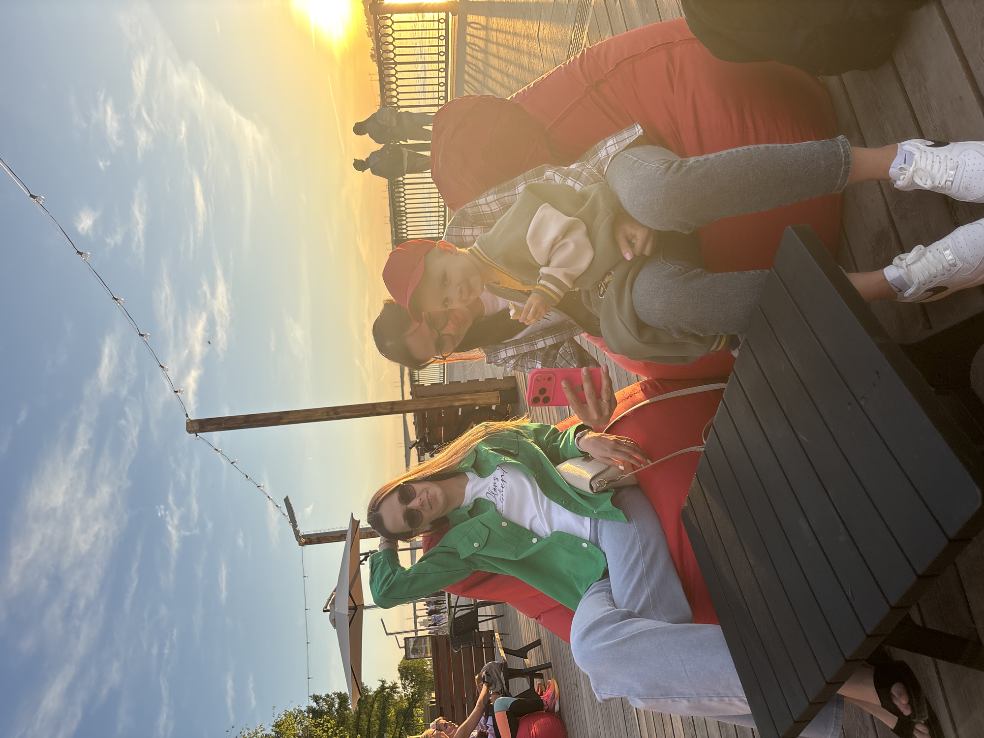 The image captures a scene on a sunny day, likely during sunset, in Perm, Russia, identifiable by the riverside setting and the presence of individuals standing on the embankment. Three people are the main subjects: a woman with long brown hair wearing a green jacket and sunglasses, holding a beige purse; another woman taking a picture with a pink smartphone, possibly of the third subject, a small child wearing a red baseball cap, seated on the lap of the second woman. They are positioned on red beanbag chairs, seated beside a small, dark-colored, square table on a wooden deck. The background shows the Kama River embankment. String lights are hung above the deck area.