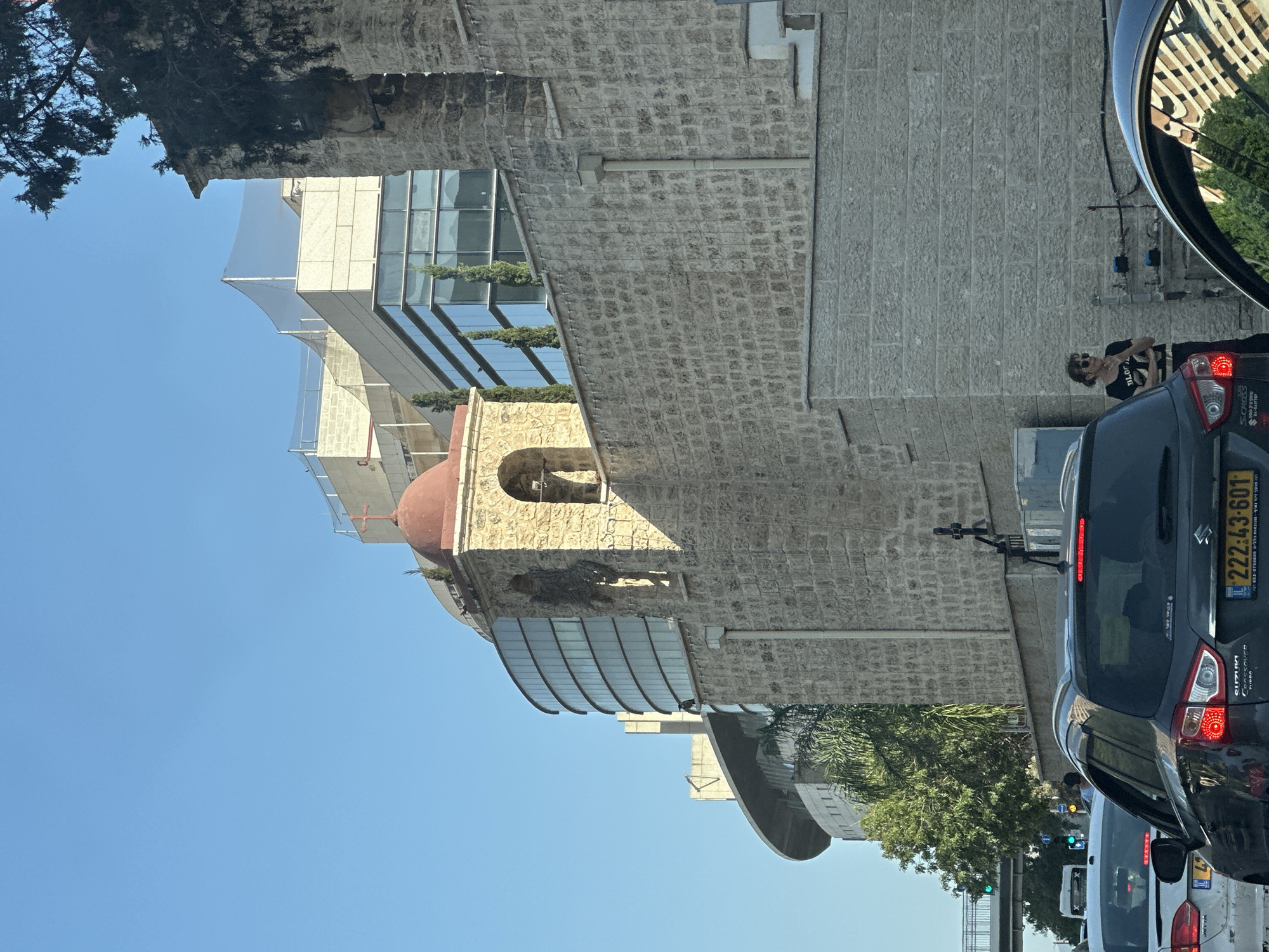 The image captures a street scene in Haifa, Israel, dominated by a stone building with a red-domed bell tower topped by a cross. Adjacent to this historical structure is a modern, multi-story building with a curved, glass facade, reflecting the sky and surrounding environment. In the foreground, traffic is visible, with several cars, including a dark gray Suzuki Crossover with Israeli license plates (222-43-601), stopped on the road. A person, a woman with short, wavy hair and sunglasses, stands near the stone wall, wearing a black t-shirt and dark pants. The sky is clear and blue, indicating daytime. Trees and other vegetation are present, with a prominent green tree on the left side of the image. A pedestrian bridge can be seen in the distance, with green traffic lights visible on a street below. The overall impression is a juxtaposition of old and new architectural styles within an urban environment.