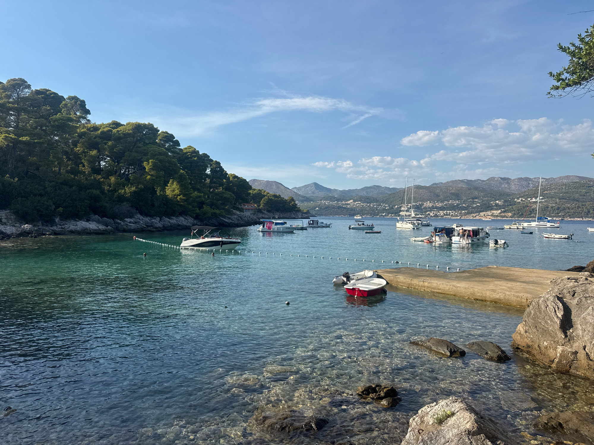 This image depicts a coastal scene in Dubrovnik, Croatia, likely within a sheltered bay or harbor. The water is remarkably clear, revealing a rocky seabed. Several boats are moored in the bay, ranging from small motorboats, some with people on board, to larger sailboats. A concrete pier extends into the water from the right side of the frame, with a small red and white boat tied nearby. Lush green trees and vegetation cover the hillside that slopes down to the water's edge on the left. In the distance, hills and mountains, possibly part of the Dalmatian hinterland, rise against a clear blue sky with scattered white clouds. The overall impression is one of tranquility and a beautiful Mediterranean summer day.