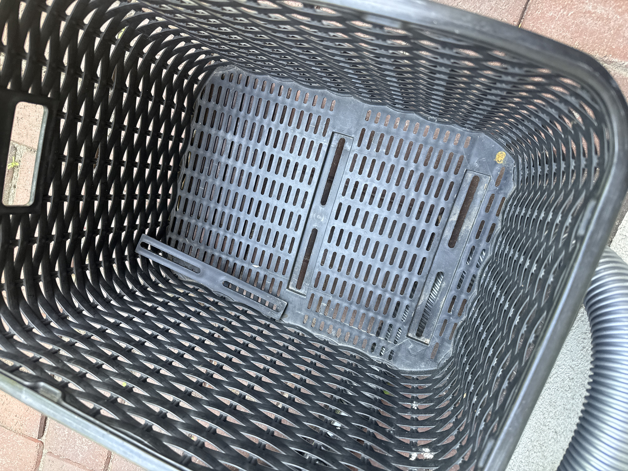 This is a close-up, overhead shot of the inside of a black plastic shopping basket. The basket has a perforated design with a woven pattern on the sides and a grid of rectangular holes on the bottom. There are a few visible dirt marks on the interior surfaces. A gray, corrugated hose is visible along the right edge of the basket. The basket is placed on a surface that appears to be made of brick pavers. There is no text visible in the image. The lighting suggests it is daytime, but the weather is not discernible. The scene is still, with no activity taking place.