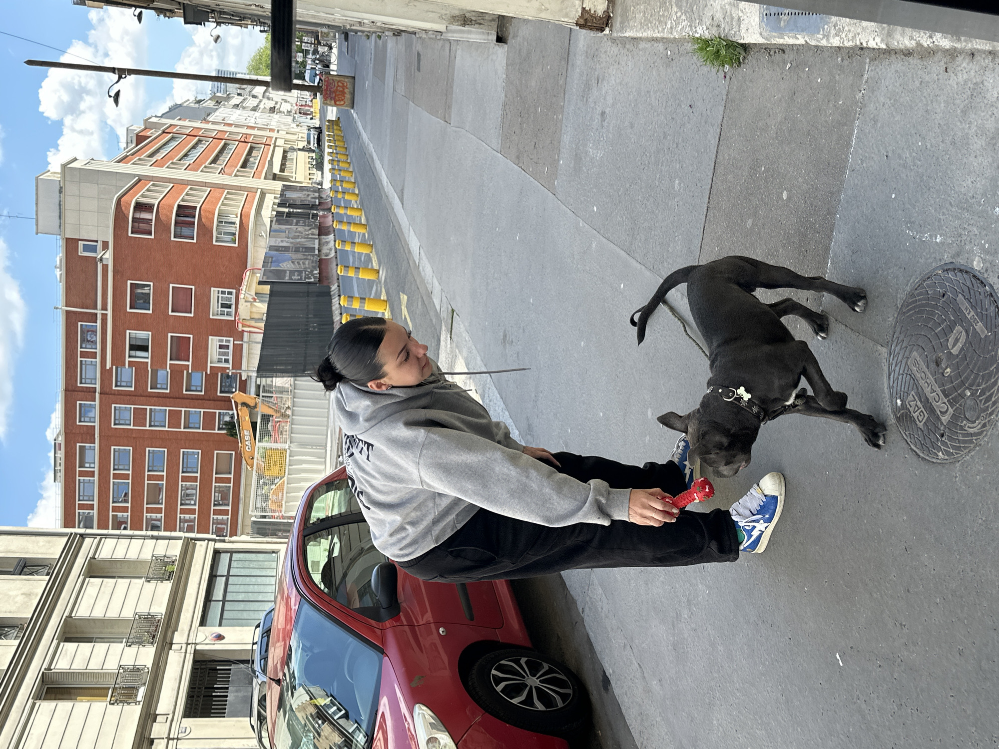 The image captures a woman standing on a Parisian sidewalk with a black dog. The woman, wearing a grey hoodie and black pants, is holding a red dog toy. The dog is positioned beside her, looking up at the toy. They are standing next to a red car parked along the curb. In the background, Parisian buildings can be seen under a partly cloudy sky. Yellow bollards are lined along the road. A manhole cover is visible on the sidewalk near the dog. Construction is present.