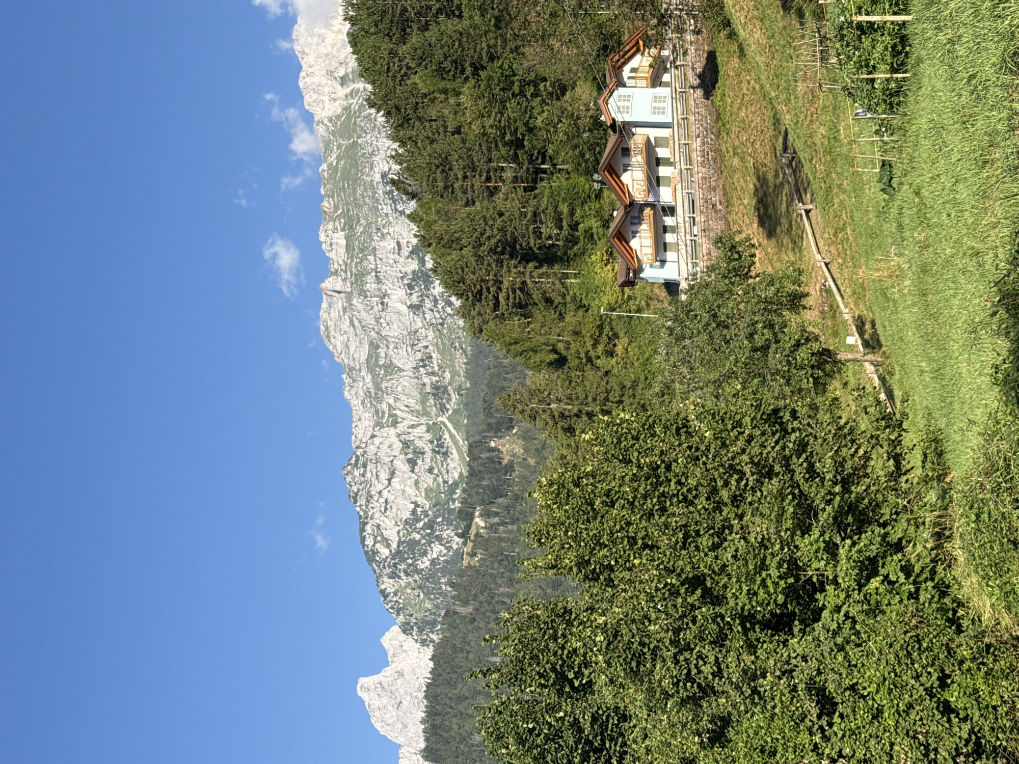 In the image, a clear blue sky stretches above a majestic mountain range with rugged, rocky peaks and green forested slopes. In the mid-ground, nestled on a hillside, are three light blue houses with white trim and brown roofs. The houses have multiple balconies with wooden railings. A stone retaining wall supports the houses, and a sloped grassy yard with some vegetation is visible in the foreground. The overall scene depicts a tranquil mountain landscape with residential structures. The provided information indicates the location is in the city of Molveno, Italy.