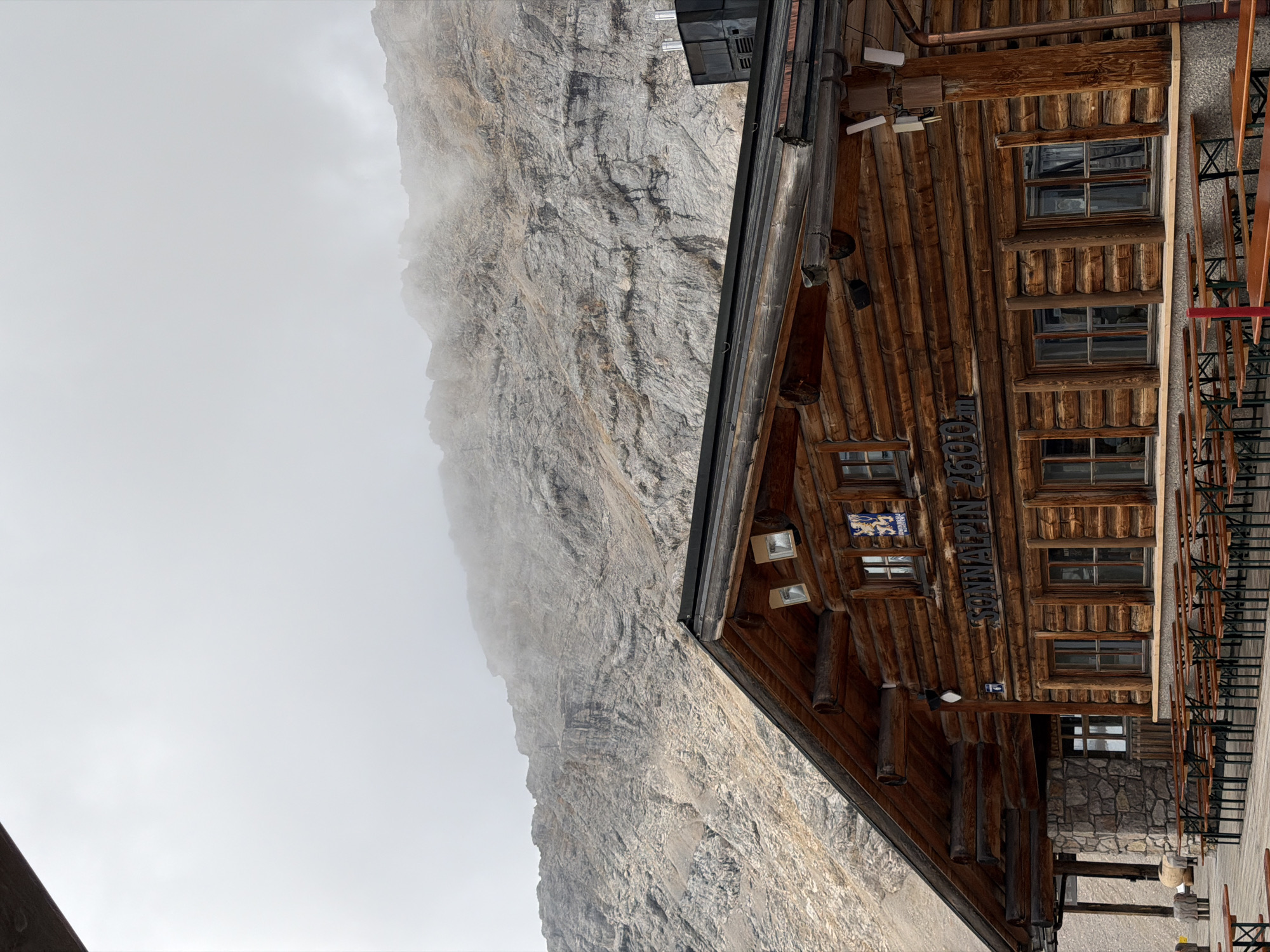 The image shows a wooden lodge building with "SONNALPIN 2600m" carved into its facade. The lodge is situated in a mountainous, possibly alpine, environment. The mountain face behind the lodge is rocky and partially obscured by fog or low clouds. In front of the lodge, numerous wooden picnic tables and benches are arranged on a wooden deck. The architecture of the lodge, with its log construction and stone foundation, suggests a mountain hut or restaurant. A crest with a blue background and white symbols is visible on the upper part of the lodge wall. The overall impression is of a remote, high-altitude establishment designed to accommodate visitors in a natural mountain setting, likely in the Alps, given the elevation indicated on the building. Based on the name "Sonnalpin 2600m" and its location, it is likely referring to the Sonnalpin restaurant on the Zugspitze mountain, the highest peak in Germany, near Garmisch-Partenkirchen.