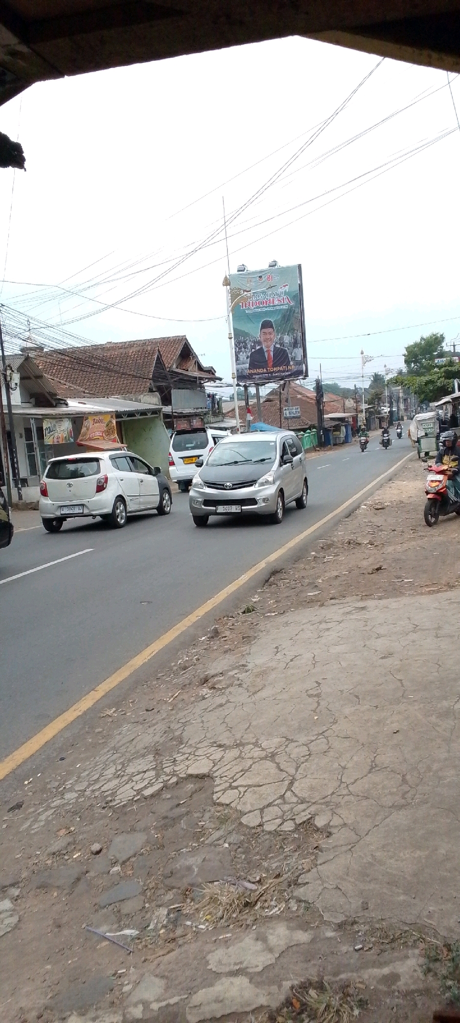 The image shows a street scene in Cilaku, Indonesia, with a billboard prominently displayed in the background. The billboard features a portrait of a man wearing a suit and tie, with text in Indonesian that appears to be political or promotional. Several cars and motorcycles are on the road, suggesting traffic activity. Buildings, including what appears to be a house with a tiled roof and a shop with signage, are visible along the roadside. The road itself is paved but shows signs of wear and tear, with cracked concrete on the sidewalk. The overall atmosphere suggests a moderately busy urban or semi-urban environment.
