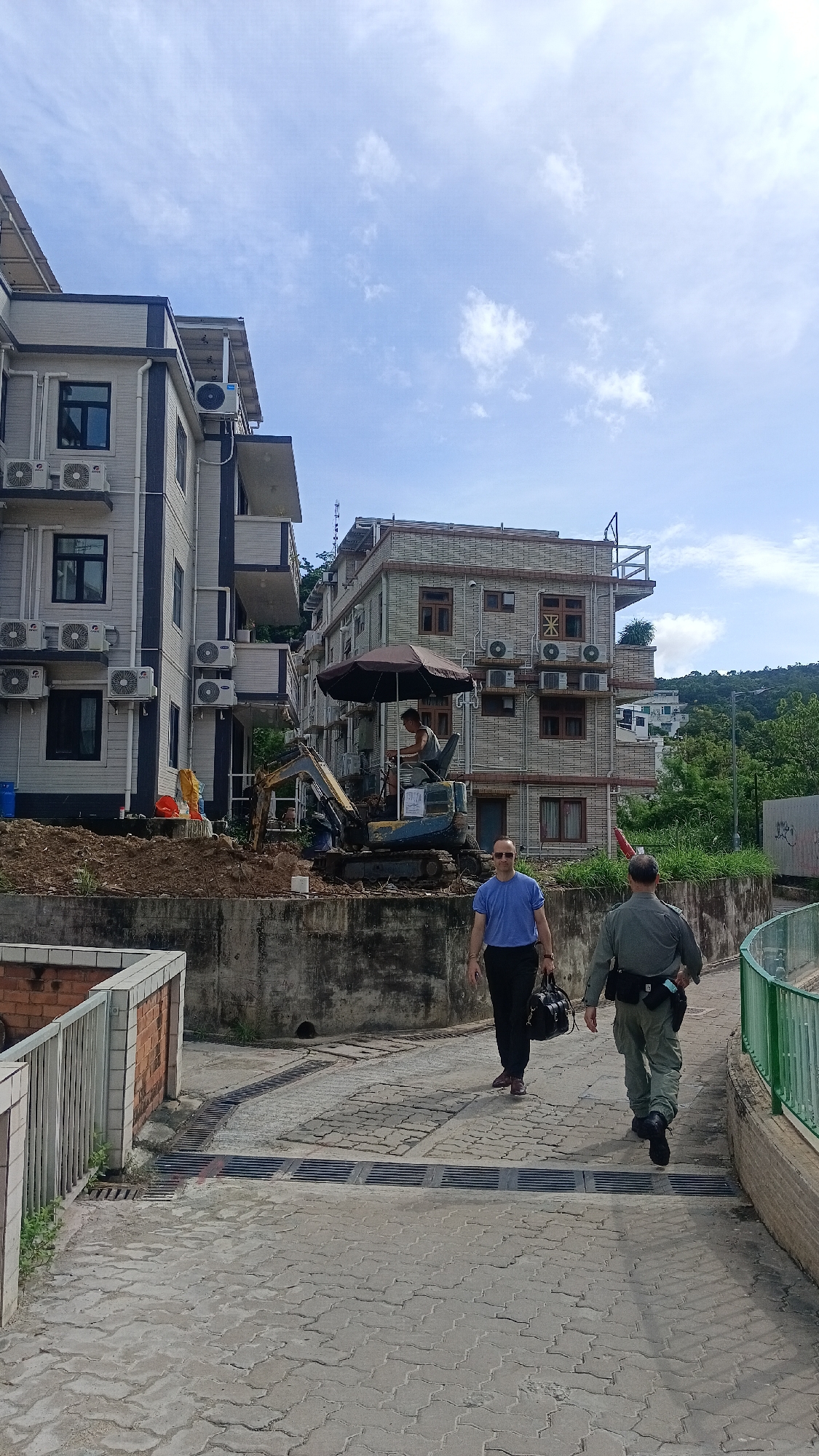 Two men walk away from the camera on a brick-paved walkway. The man on the left wears a blue t-shirt and black pants and carries a black duffel bag. The man on the right wears a green uniform with a belt and black boots. In the background, a small excavator is parked on a pile of dirt in front of a multi-story building. Another multi-story building is to the left of the excavator. The buildings are residential with numerous air conditioning units visible. A brown umbrella is open over the excavator. The sky is blue with scattered white clouds. The scene appears to be in a residential area of Hong Kong, China, possibly undergoing construction or renovation.
