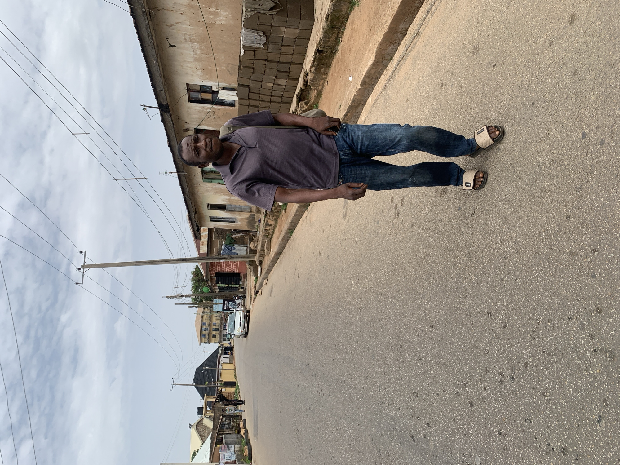 A man stands on a street in Jos, Nigeria. He is wearing a purple collared shirt, blue jeans, and sandals. A shoulder bag is slung across his chest. He faces forward, looking slightly to his left.
The street is asphalt, and there are buildings on either side. Power lines run along one side of the street. In the distance, vehicles are visible on the road. The sky is overcast.
