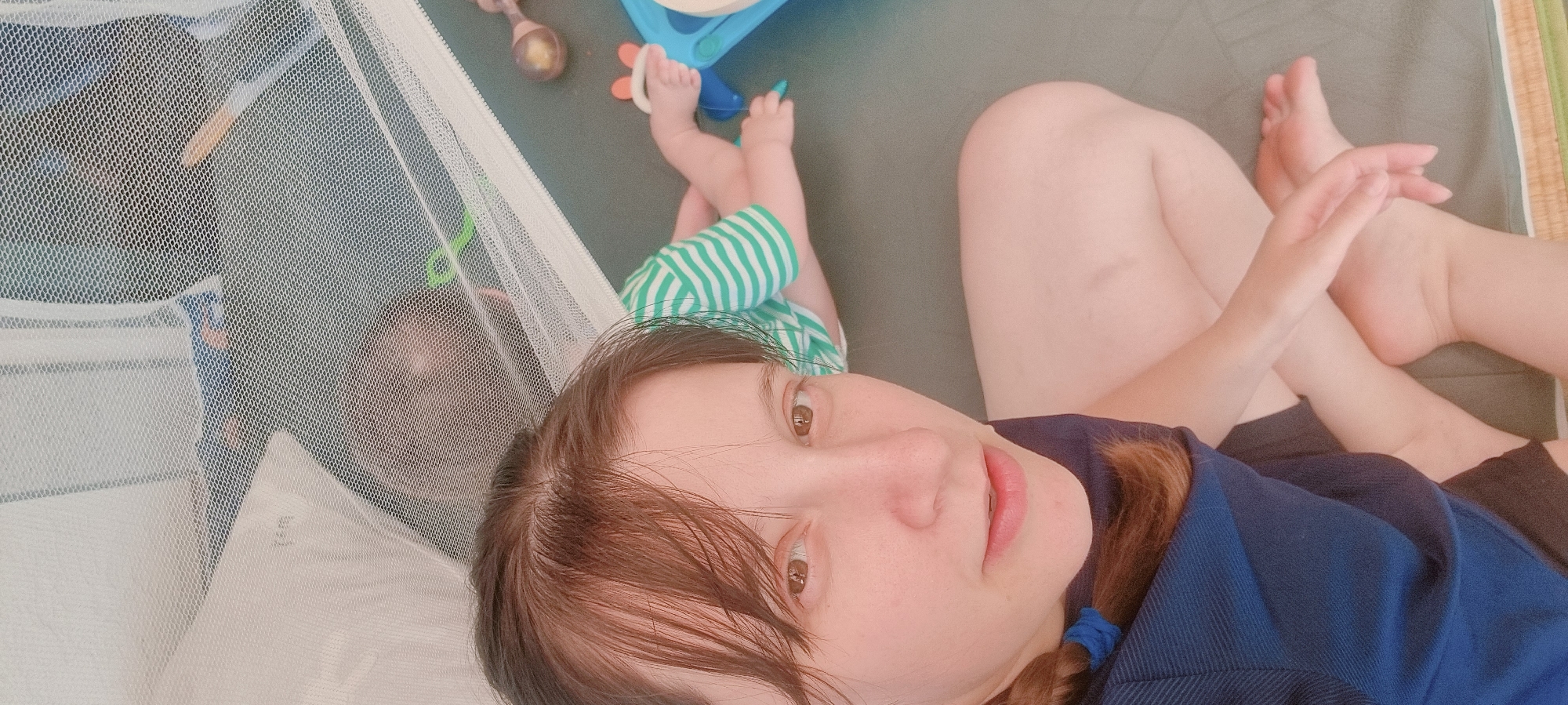 A woman lies supine inside a mosquito net enclosure. A baby wearing green and white striped clothing lies adjacent to the woman, within the netting. The woman wears a blue shirt, and her hair is partially braided. Baby feet are visible. There are various toys including a wooden rattle, and a blue and orange plastic toy. The background suggests a playmat on the ground. The location is Dunnae-myeon, South Korea.