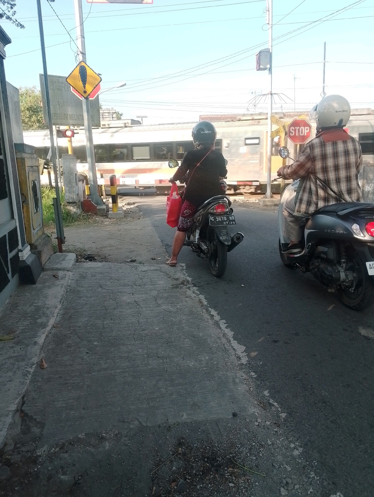 The image depicts a railway crossing in Ngebra, Indonesia, on a sunny day. Two individuals on motorcycles are stopped at the crossing, waiting for a train. The person on the left is riding a black motorcycle and is wearing a black helmet and a dark top with a red and white patterned skirt. They are holding a red bag. The person on the right is riding a dark gray scooter and is wearing a light-colored helmet and a plaid shirt. A red and white octagonal "STOP" sign is visible near the crossing. A train with orange and white stripes is passing through the crossing. The environment includes a paved road, a concrete sidewalk, grassy areas, and various signs, including a yellow diamond-shaped sign with a black exclamation mark and text in Indonesian. Overhead power lines are also visible.