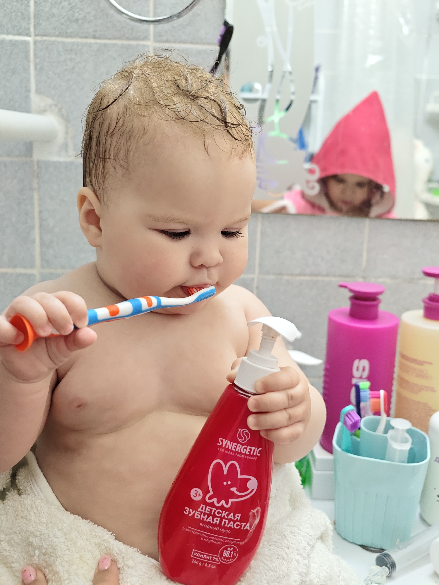 A baby, wrapped in a white towel, is in a bathroom, brushing their teeth with an orange, white, and blue toothbrush. In the baby's other hand, they are holding a red bottle of "Synergetic" children's toothpaste. The label on the bottle is in Russian and indicates it is a berry mousse flavored toothpaste for ages 3+. Behind the baby is a white countertop with various toiletries, including pink bottles, a light blue toothbrush holder, and other hygiene products. In the mirror behind the baby, there is a reflection of a child wearing a pink hooded towel. The bathroom walls are tiled in a gray color. The image was taken in Chaikovsky, Russia.