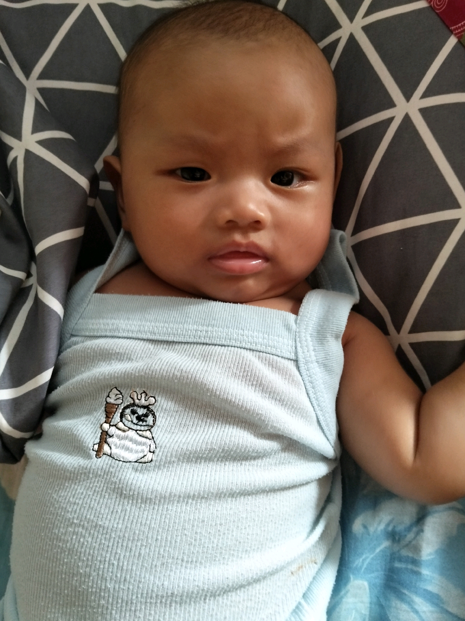 A close-up, high-angle shot shows a baby lying on their back on a patterned surface, in what appears to be a home environment in Gempolan, Indonesia. The baby, with dark skin and hair, is wearing a light blue tank top with a cartoon embroidery of a character holding an ice cream cone. The background features a grey and white geometric patterned fabric, possibly a blanket or pillow. The baby's face, particularly the eyes, nose, and mouth, is visible, with a slight furrow in the brow. The lighting is even, highlighting the textures of the baby's skin and clothing.