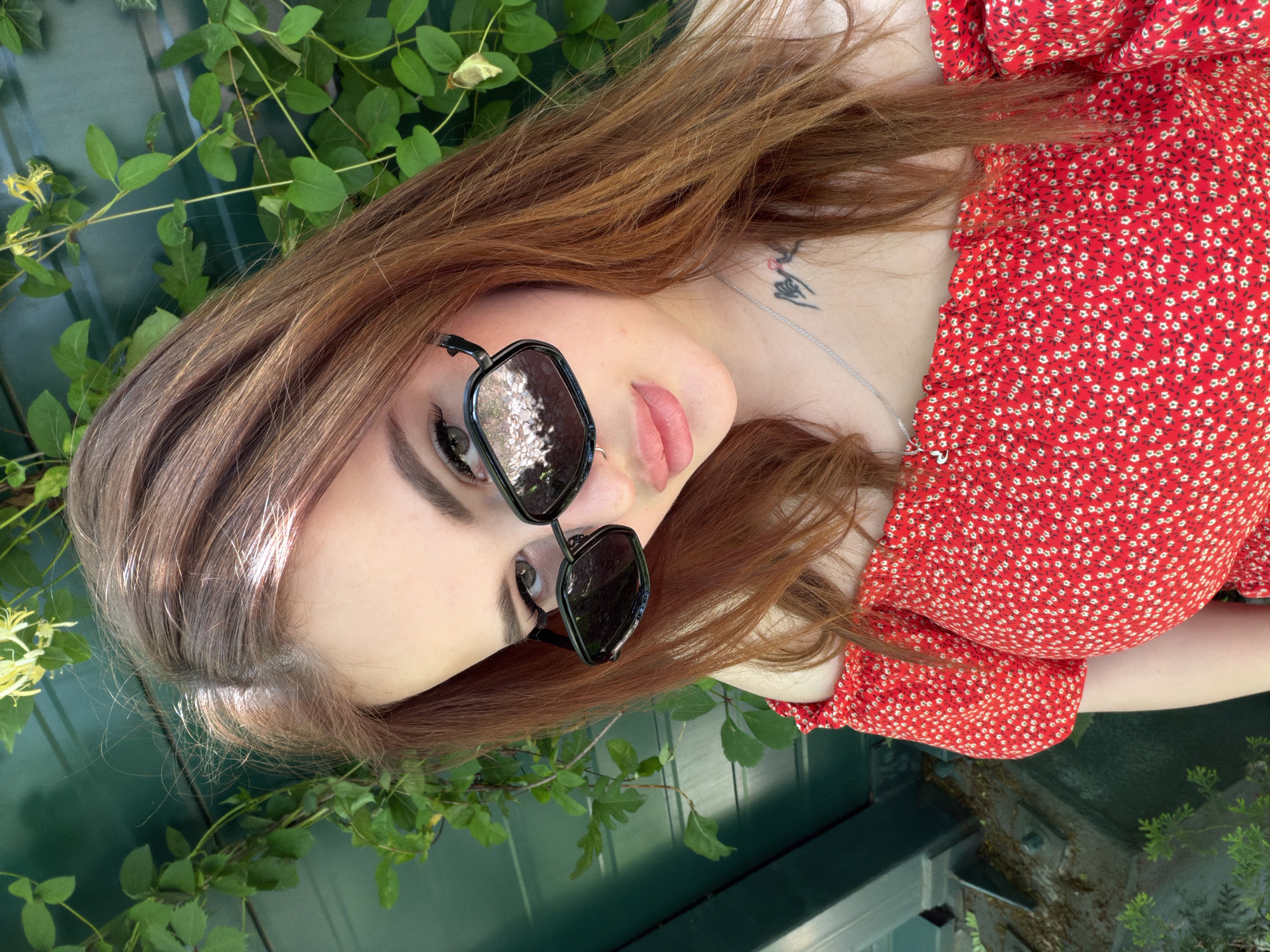 A young adult female is depicted in an outdoor, sunlit self-portrait. She has long, reddish-brown hair and is wearing black hexagonal sunglasses, which reflect the environment. A small silver hoop nose ring is visible in her left nostril, and a thin silver chain necklace with a small pendant adorns her neck. On her right clavicle, a dark-ink tattoo, appearing to be stylized script or a symbol incorporating a small red heart, is present. She is dressed in a red off-the-shoulder top featuring a repeating pattern of small white flowers with light green centers. Her lips appear glossy. The background consists of a dark green paneled fence or wall, largely covered by lush green foliage, including vines with visible yellow honeysuckle flowers. Strong direct sunlight illuminates the scene. To the lower left, a dark metallic structural element, possibly a beam or railing, with minor debris, is partially visible. The image contains no discernible visual markers to confirm the specified location as Krasnodar, Russia.