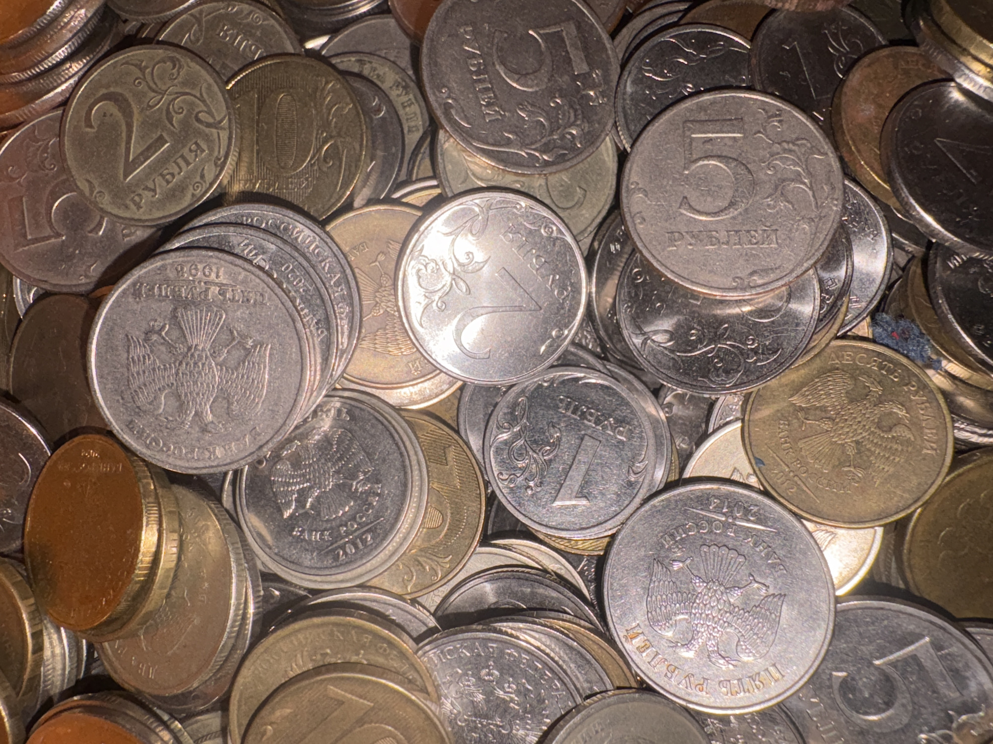 The image displays a close-up, overhead view of a large pile of Russian coins, primarily rubles. The coins are of various denominations, including 1, 2, 5, and potentially others, indicated by the visible numerals and inscriptions. Some coins are silver-colored, while others are gold or copper-toned. The coins are stacked and jumbled together, with some overlapping others. The lighting creates shadows and highlights that emphasize the texture and details of the coins, such as the double-headed eagle emblem and Cyrillic lettering. There are no people, animals, or any specific environmental features visible beyond the coins themselves. The context of Georgiyevsk, Russia is not visually represented in the image.