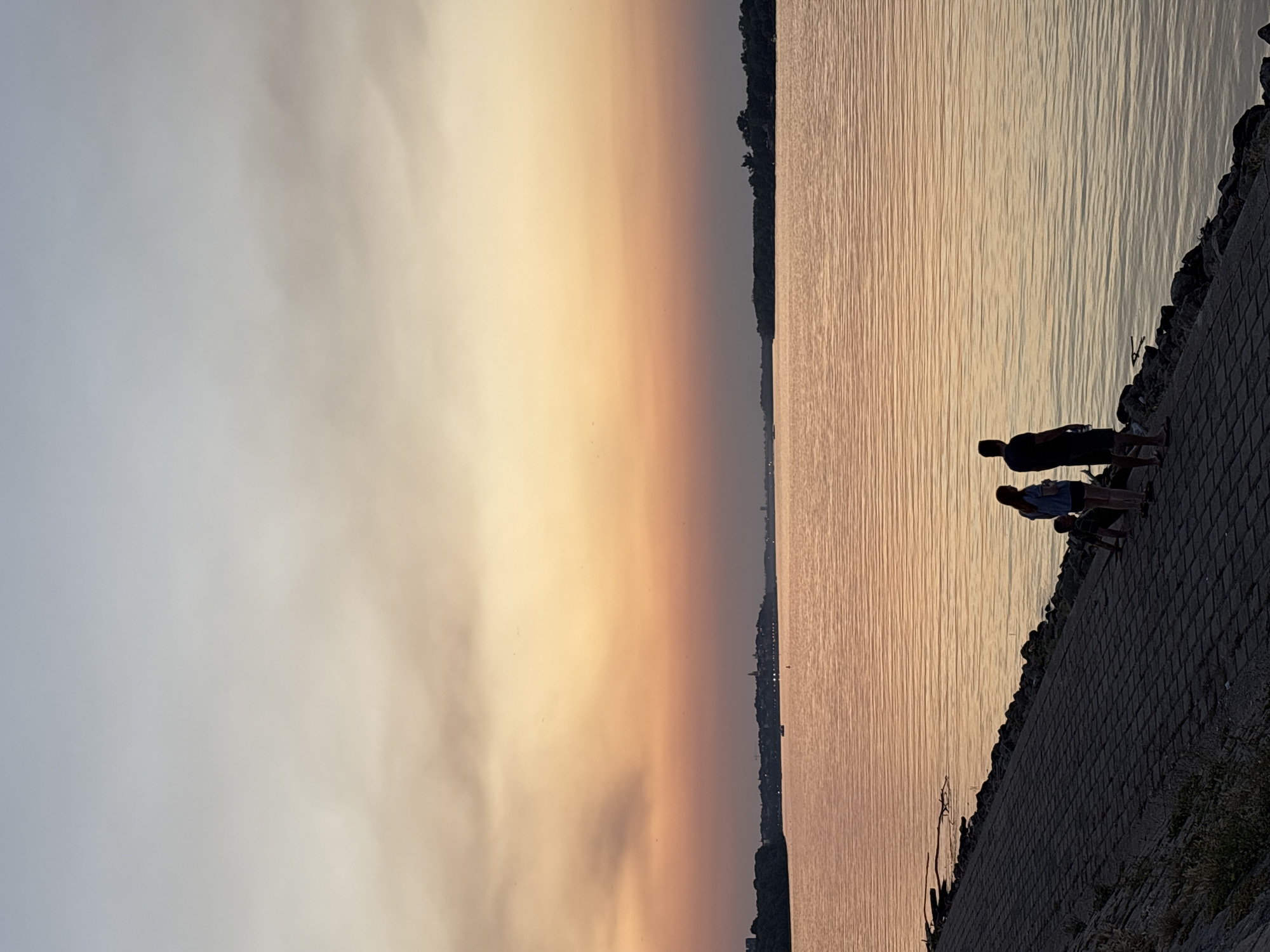 A vertical, wide shot captures the sunset over the Danube River in Belgrade, Serbia. The sky displays a gradient of colors, transitioning from a pale blue at the top to warm orange and pink hues near the horizon. The river reflects these colors, creating a shimmering surface.
In the foreground, a paved walkway slopes down to the water's edge, lined with rocks. Silhouetted against the sunset are three figures: a woman with long hair and a child standing beside her, and a man standing slightly apart from them. The woman is wearing a light-colored top and dark shorts. The child is a smaller figure, positioned between the woman and the man. The man is wearing dark shorts and a dark t-shirt. They appear to be observing the sunset or the river.
Across the river, the opposite bank is visible as a dark, silhouetted line of trees and buildings, hinting at a cityscape. A few small boats are present on the water. The overall atmosphere is serene and tranquil, characteristic of a peaceful evening by the river.