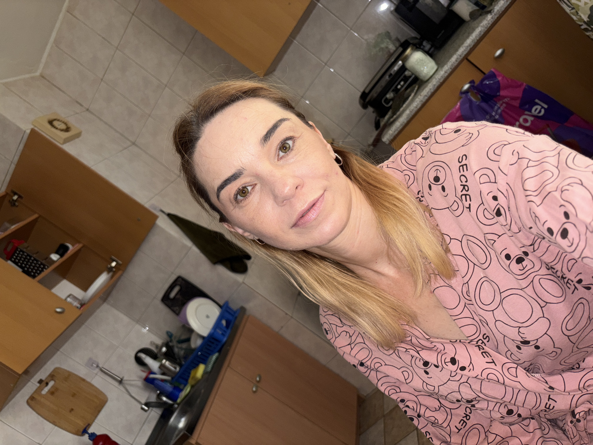 A selfie is taken in a kitchen, likely in Saint Petersburg, Russia, as indicated by a visible shopping bag from a Russian store. The subject, a woman with blonde hair and brown eyes, is wearing a pink pajama top with a teddy bear pattern. She is positioned in the foreground, with a kitchen sink and cabinets visible in the background. The kitchen appears to be functional, with dishes in a drying rack and various appliances on the counter. The lighting suggests it is indoors, possibly at night or in a dimly lit room.