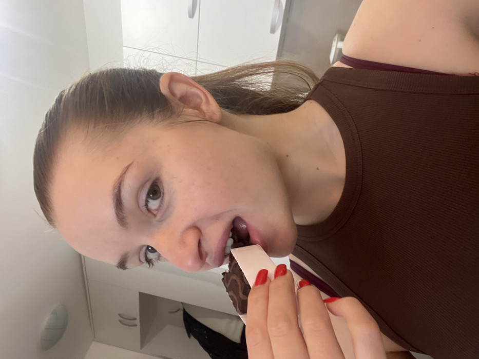 A young woman with light skin, brown hair pulled back in a ponytail, and wearing a brown tank top is shown eating from a box. She holds the box with red manicured nails and appears to be looking directly at the camera. Her expression is one of enjoyment. The background includes a white cabinet and part of a garment. The image is slightly rotated.