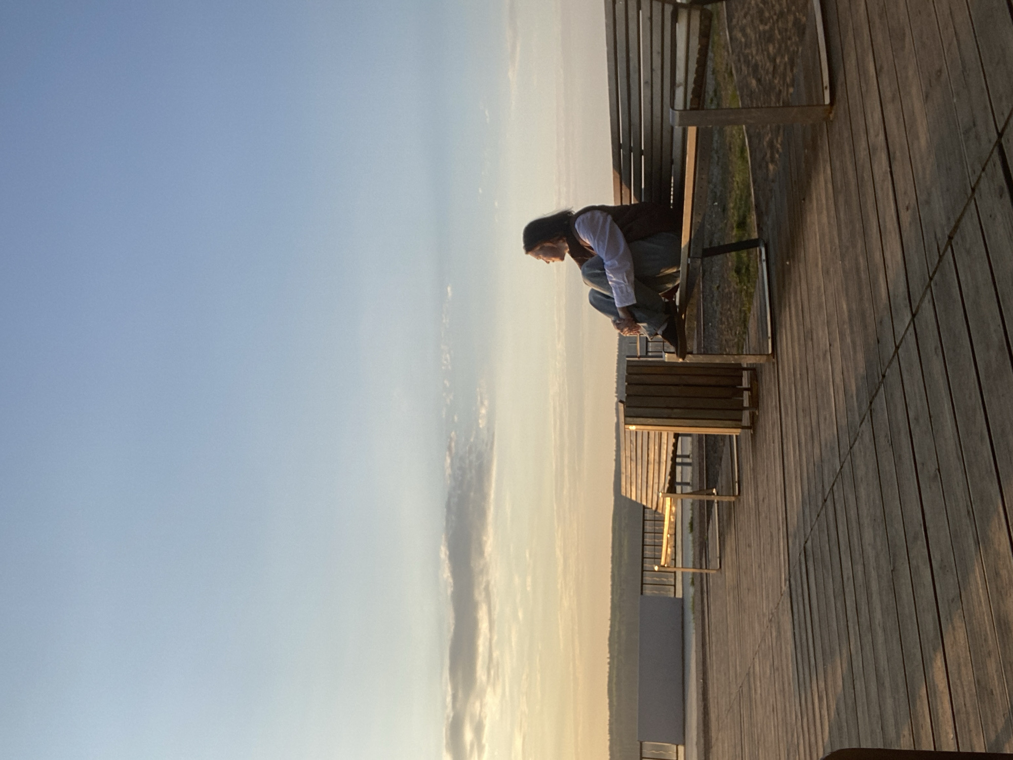 A young adult sits on a wooden bench on a wooden boardwalk or deck, looking down and touching or adjusting their shoes. The person is wearing a white long-sleeved shirt, a dark vest, jeans, and a dark hat or head covering. The boardwalk is adjacent to a grassy area with vegetation. In the distance, there is a tall building or structure. The sky is a gradient of light blue to pale yellow, suggesting either sunrise or sunset. The location is Chaikovsky, Russia.