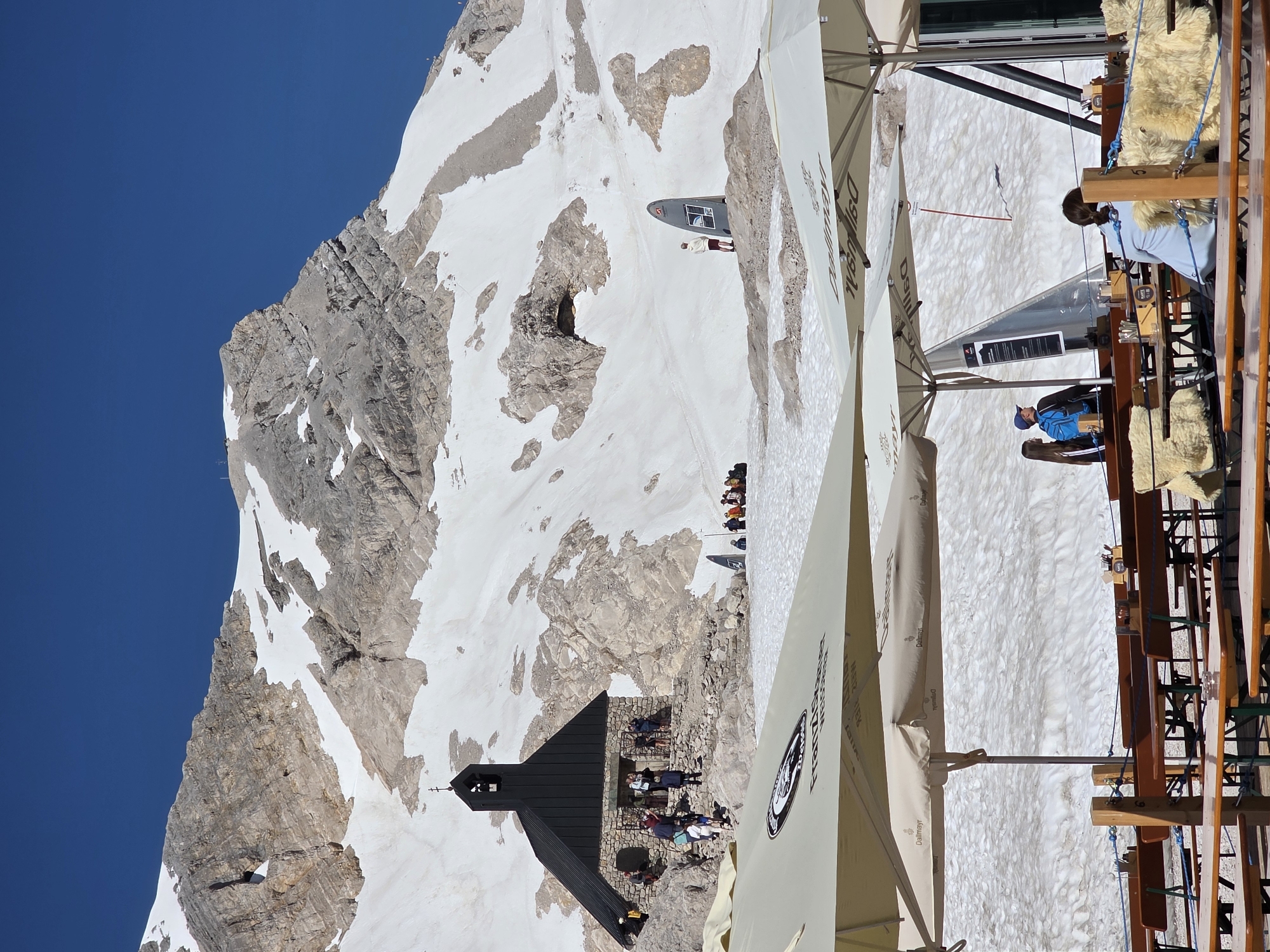 This is a vertical, eye-level, medium-wide shot of the Zugspitze summit in Garmisch-Partenkirchen, Germany. In the middle ground, a small stone chapel with a black roof and steeple stands on a rocky outcrop against a backdrop of a snow-covered mountain. Several people are visible on and around the chapel. In the foreground, two large, cream-colored umbrellas with logos are angled into the frame, casting shadows on an outdoor seating area with wooden tables and benches. The ground in the foreground and middle ground is covered in snow. In the background, the vast, rocky, snow-capped Zugspitze mountain range under a clear blue sky dominates the scene.