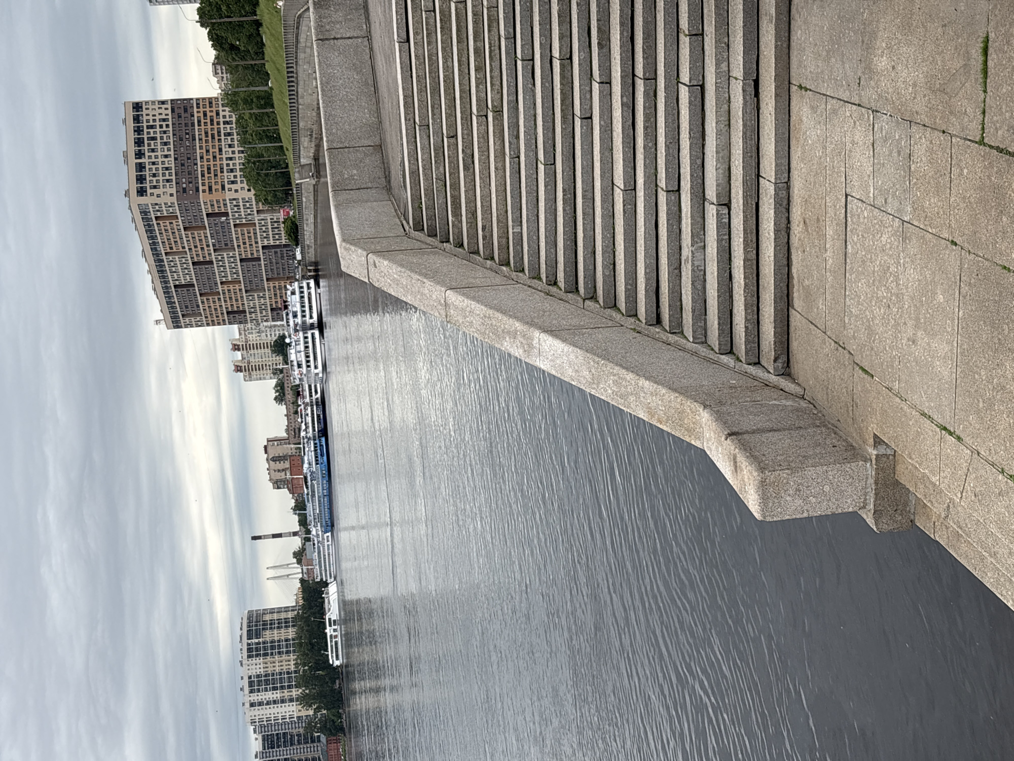 The image shows a view of the Neva River in Saint Petersburg, Russia, on a cloudy day. In the foreground, there is a wide stone staircase leading down towards the water. The river itself is calm, with several large passenger ships docked along the opposite bank. Beyond the ships, a cityscape with modern apartment buildings and a distinctive, irregularly shaped building is visible. Trees line the embankment, and a bridge can be seen in the distance. The sky is overcast, casting a diffused light over the scene. There are no people visible in the foreground or midground, though a single person is faintly visible walking on the embankment in the far right distance.