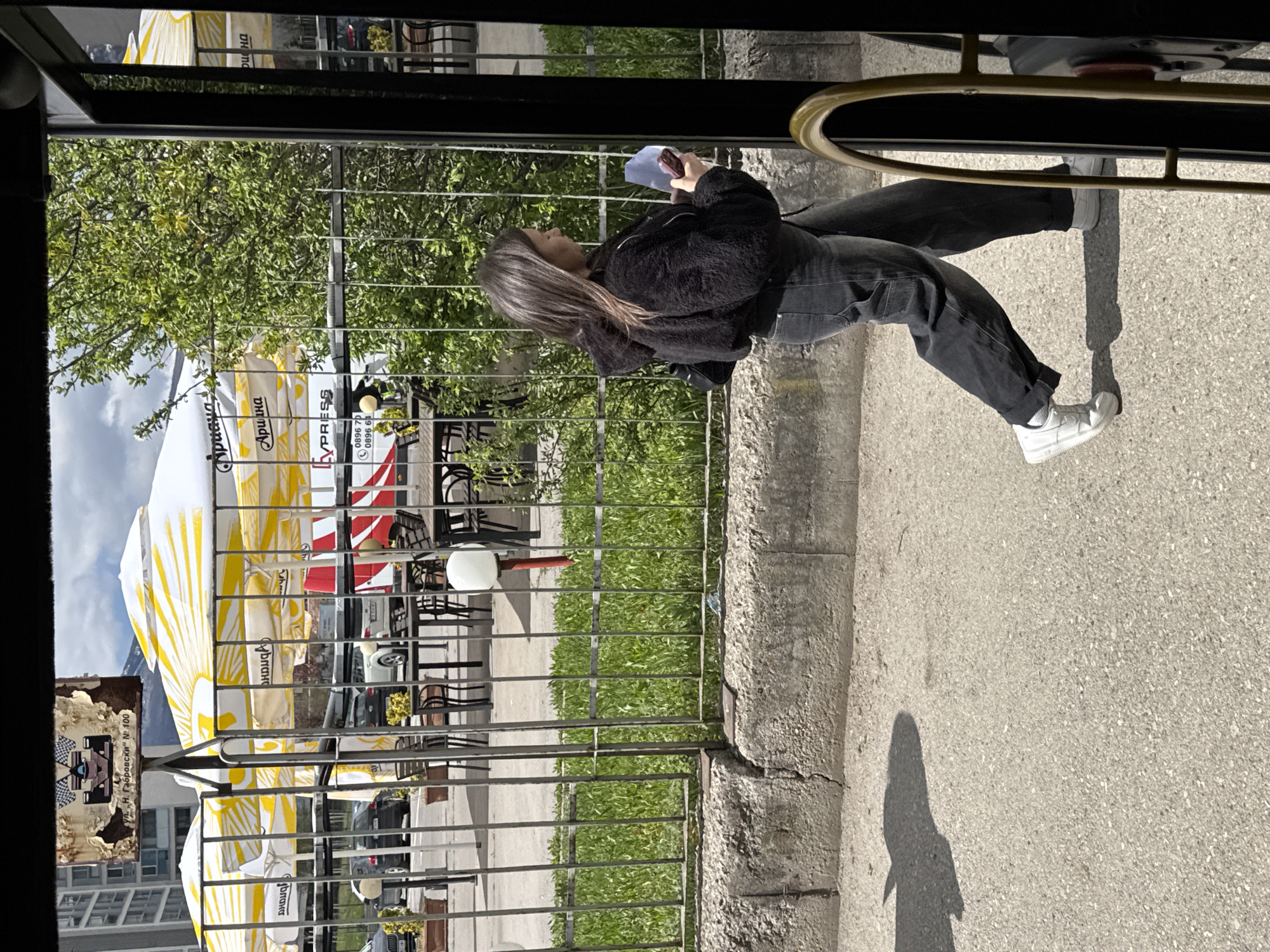 This media file captures an outdoor scene on a sunny day, likely taken from inside a vehicle or structure looking outwards. The image is rotated 90 degrees clockwise.
The central subject is a young woman standing in profile, viewed from the waist up. She has long, straight dark hair and is wearing a fuzzy black jacket, dark pants, and white sneakers. She is holding a light blue object, possibly a phone, in her hands and appears to be looking at it. Behind her is a metal fence or railing, and beyond that, lush green trees.
In the background, there is an outdoor cafe or seating area with several large white and yellow striped parasols or umbrellas, and visible tables and chairs. Cars can be seen parked or driving in the distance. The ground is paved, appearing to be an asphalt or concrete surface, and a distinct shadow of a person (likely the photographer) is cast on it.
Notable details include:
- **Time of day:** Bright daylight, indicated by strong, well-defined shadows.
- **Weather:** Sunny with a clear sky visible between the trees and umbrellas.
- **Perspective:** The shot is taken from a slightly elevated position, looking down, through a dark frame and a gold/brass bar.
Visible text provides location cues:
- On the white and yellow umbrellas, the brand names "Ариана" (Ariana) and "CYPRESS" are visible, along with phone numbers "0896 70" and "0896 6". Another word "Δρασμα" (Drasma) is also on an umbrella, and "вланат" (vlanat) on another.
- A rusty sign in the background displays "ул. „Н. Габровски" № 100" (ul. "N. Gabrovski" No. 100), indicating a street address.
The scene depicts a casual, bright day in what is identified as Sofia, Bulgaria, suggesting an everyday street or cafe environment.