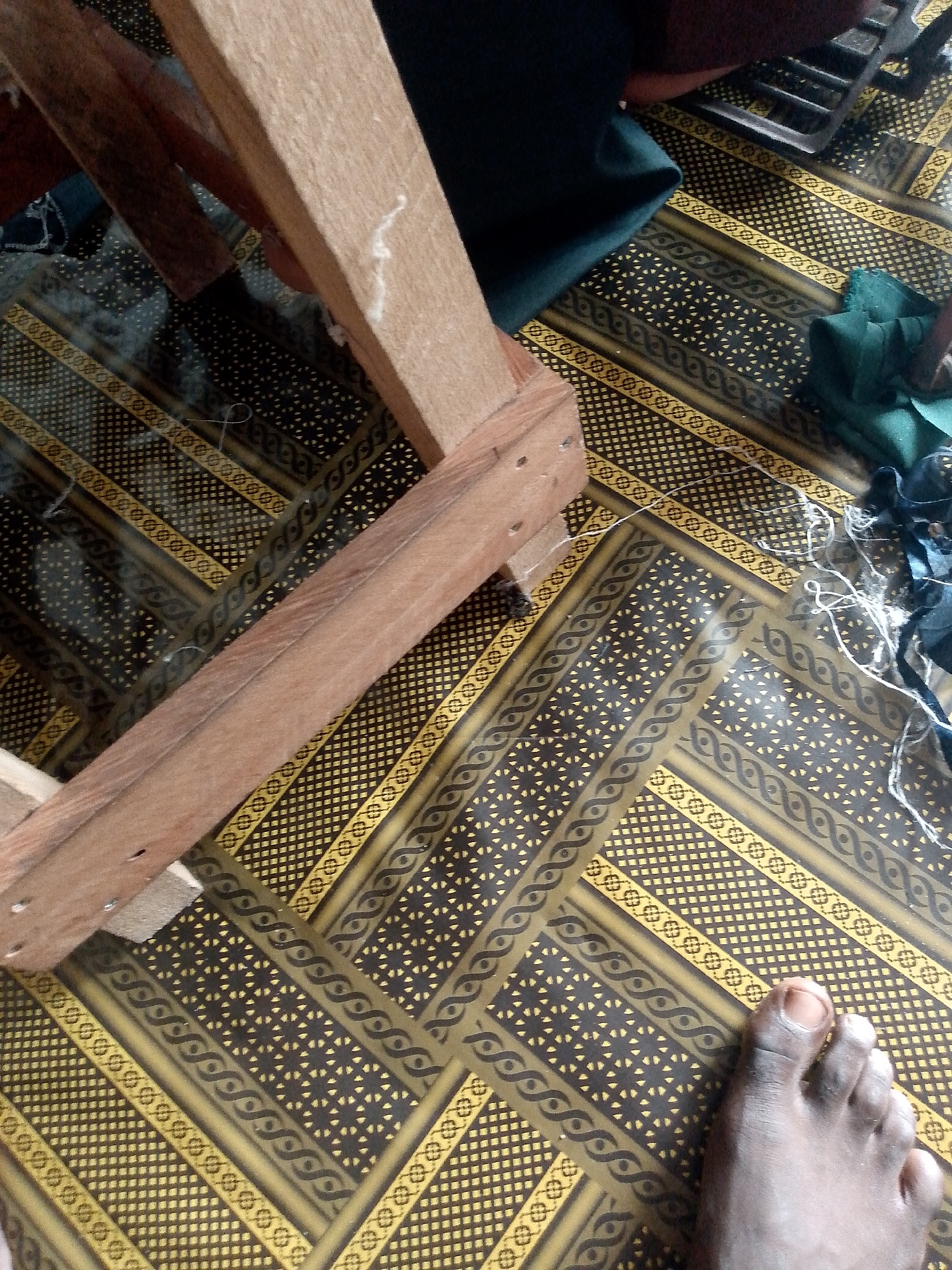 A close-up, ground-level shot shows a wooden structure resting on a patterned mat. The mat features a repeating geometric design with dark brown and yellow elements. A person's foot, with dark skin and painted toenails, is visible in the lower right corner of the frame, partially obscuring the mat. Several pieces of dark green fabric are scattered on the mat, along with white threads and a dark object that may be a sewing machine. The lighting suggests an indoor setting, possibly a workshop or a home craft area. The scene is located in Jauro Yinu, Nigeria.
