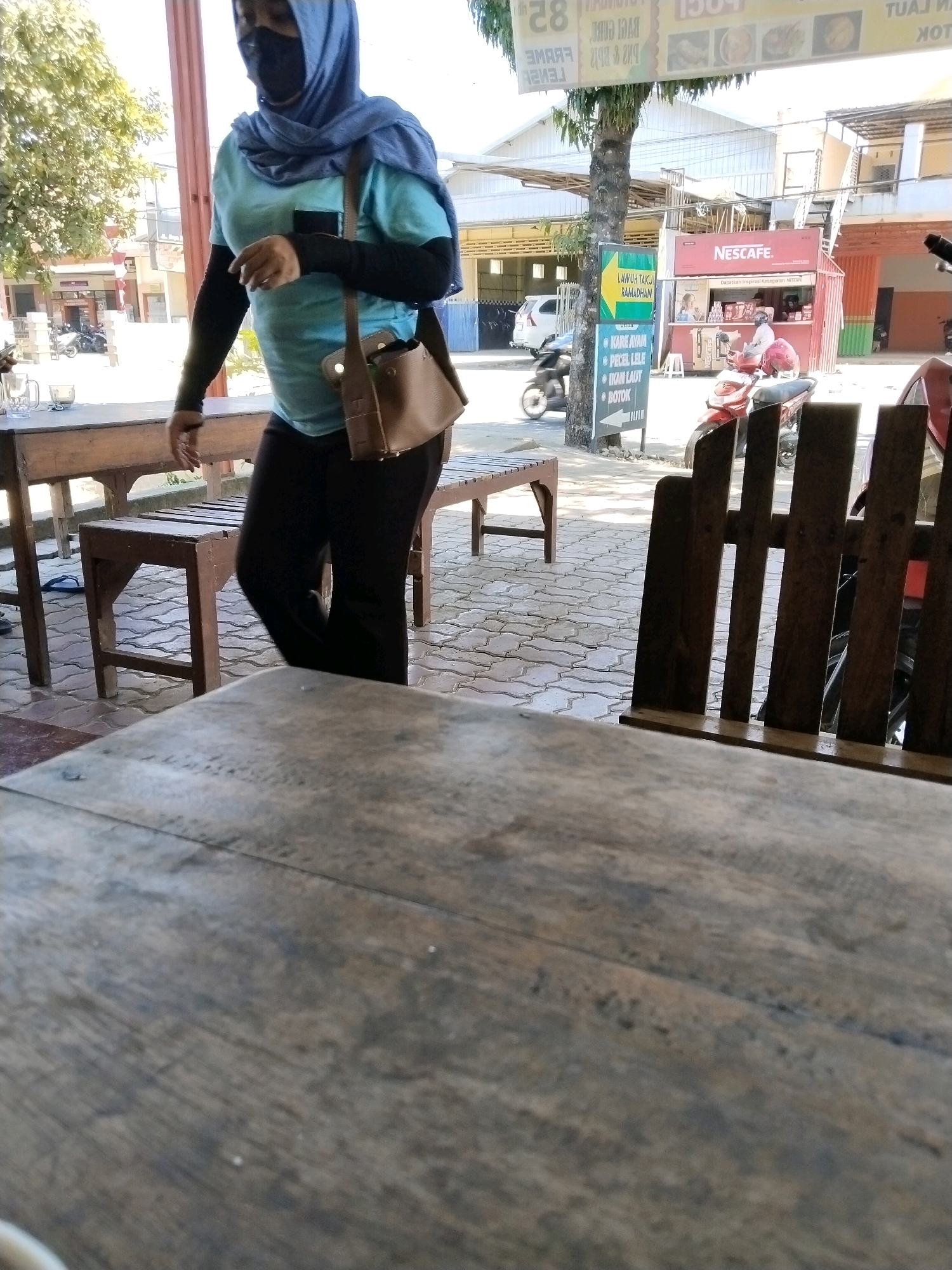 The image depicts an outdoor scene in Mergayu, Indonesia, likely a roadside dining area. In the foreground, a weathered wooden table is partially visible. Behind it, a woman wearing a blue hijab, a light blue t-shirt, and black pants walks past. She has a brown shoulder bag and is wearing black arm coverings. To her left, a wooden table and bench are set up. In the mid-ground, a sign displays "LAWUH TAKJIL RAMADHAN" and lists food options like "KARE AYAM," "PECEL LELE," and "IKAN LAUT." A red "NESCAFE" kiosk is situated behind the sign, with a person inside serving customers. Several motorcycles are parked nearby, including one in motion. The ground is paved with interlocking bricks. The background shows buildings and trees, suggesting a commercial or residential area. The lighting indicates it is daytime, with sunlight casting shadows.