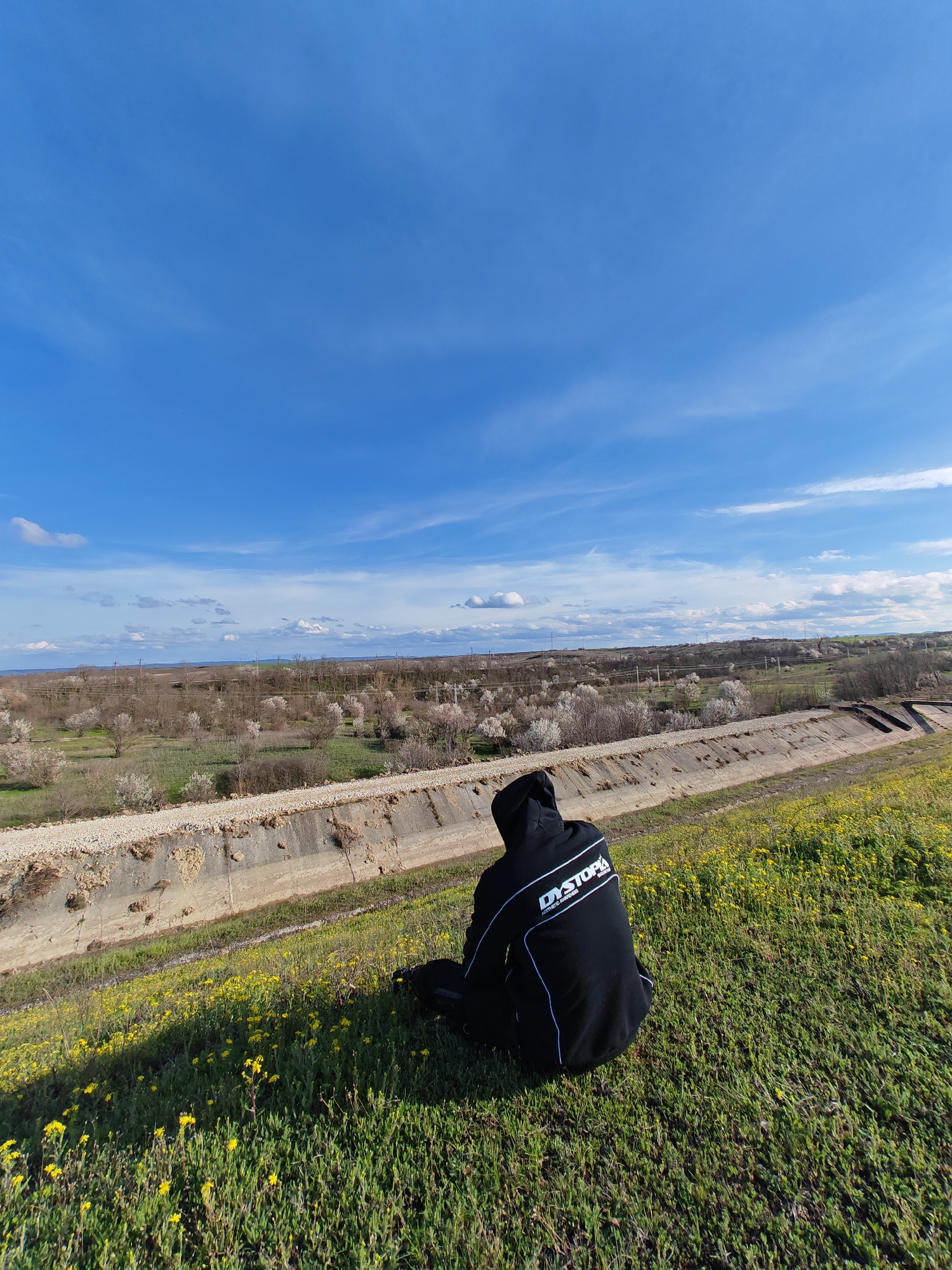 The image shows a person sitting on a grassy hill in what appears to be a rural setting on the outskirts of a populated area. The person is wearing a black hoodie with white lettering "DYSTOPIA" on the back. A concrete irrigation canal runs between the grassy hill and an agricultural landscape of trees and fields. The sky is blue with scattered clouds. Based on visual cues of landscape and infrastructure, it is consistent with the region surrounding Shumen, Bulgaria, but this cannot be determined with complete certainty. The person is facing away from the camera, seemingly contemplating the landscape. Yellow wildflowers are scattered throughout the green grass on the hill.