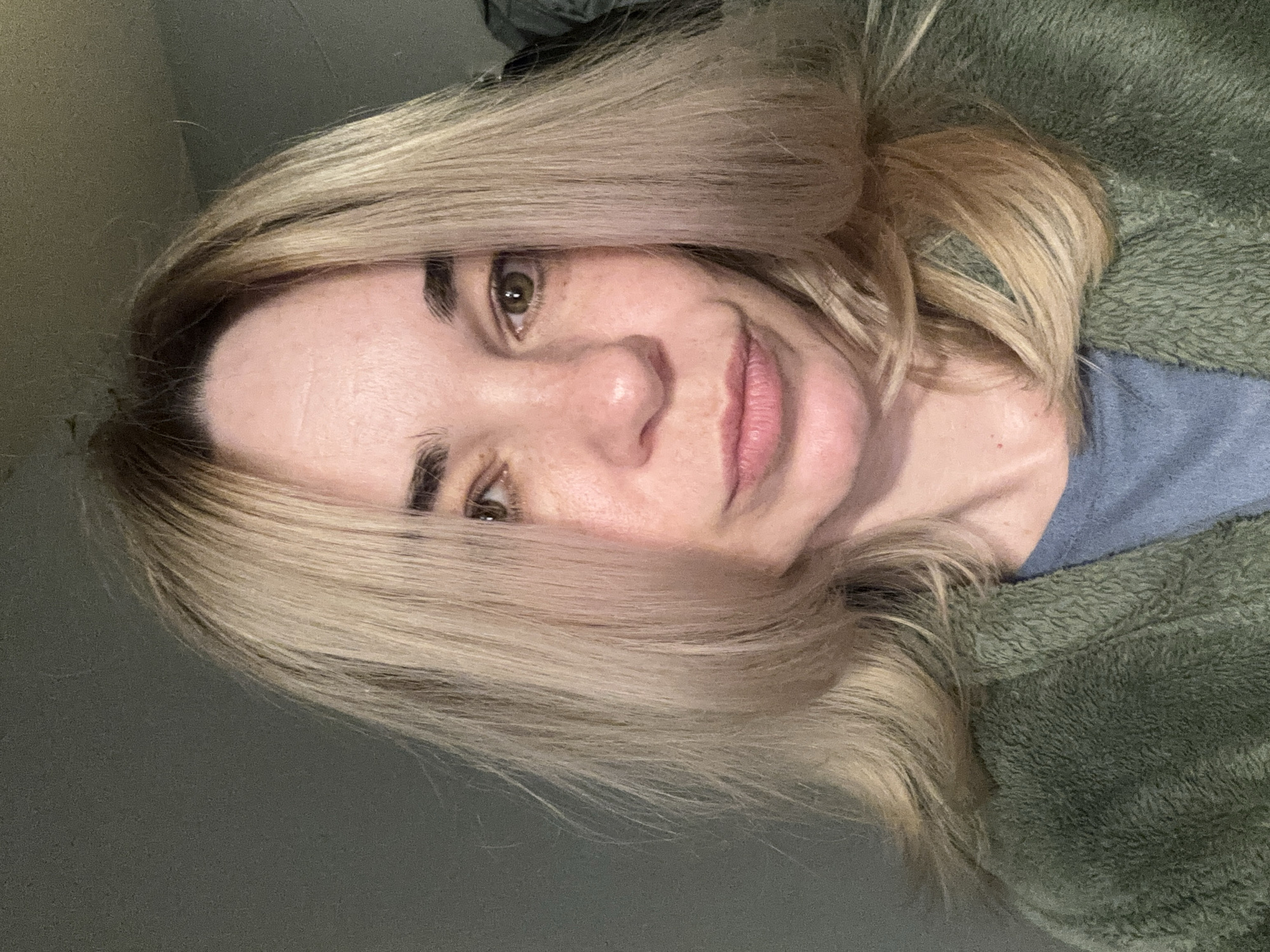 A close-up portrait of a woman with shoulder-length blonde hair. She has fair skin, brown eyes, and appears to be smiling slightly. She is wearing a green, textured jacket or sweater over a blue shirt. The background is a plain gray wall. There is no indication of the city of Vsevolozhsk, Russia, or any other location information visible in the image.