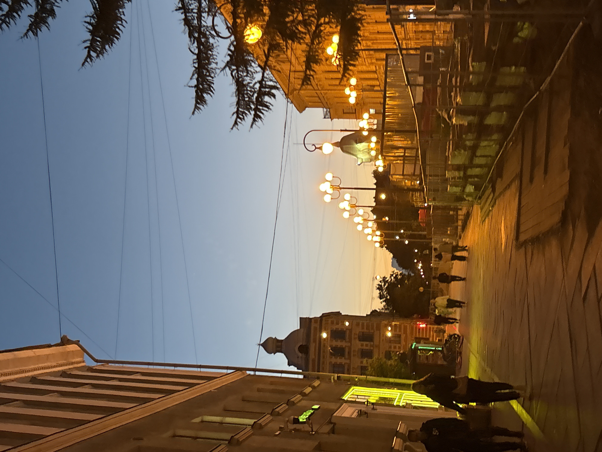 This is a street-level, eye-level, medium shot photograph taken during dusk on a street in Saint Petersburg, Russia. The sky is a deep blue, transitioning to a warm orange hue near the horizon. The light suggests it is twilight, with streetlights beginning to illuminate the scene.
On the left side of the frame, a building with a light-colored facade and a series of evenly spaced vertical windows dominates the foreground. The building's architecture appears old, with decorative elements visible near the roofline. A drainpipe runs vertically down the side of the building. Some illuminated storefronts are visible at street level.
In the mid-ground, several people are walking along a paved sidewalk. Most are walking away from the camera, towards the right side of the frame. They are dressed in casual clothing, with one person in the foreground on the left wearing a dark jacket and another woman in a dark jacket and light-colored pants walking beside them.
Further down the street, more people are visible walking. The sidewalk is lined with streetlights, which are ornate and feature multiple globes, casting a warm, yellow glow on the surroundings. Some of these lights are on, and others appear to be off.
On the right side of the frame, another building with a similarly aged architectural style is visible. This building is illuminated by the warm light from the streetlights. A prominent feature on this side of the street is a series of decorative streetlights and what appears to be a construction or renovation site, cordoned off by a fence. Stacked materials, possibly construction supplies wrapped in yellow plastic, are visible behind the fence.
A tall evergreen tree with dark needles is visible in the upper right corner of the image, partially obscuring the sky and the building behind it. Electrical wires are strung across the sky, a common sight in urban environments. The overall atmosphere is calm and urban, capturing a moment of daily life in the city as evening sets in. The specific location is suggested to be Saint Petersburg, Russia, based on the architectural style and the presence of overhead wires.