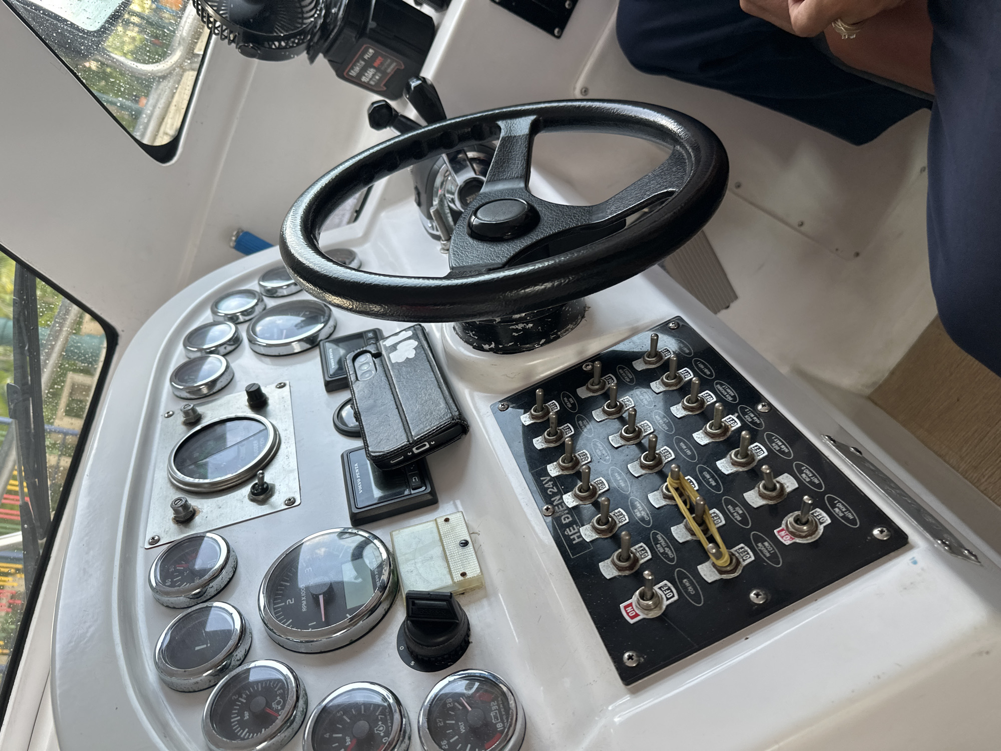 The image presents the interior helm station of a boat, located in Nha Trang Ward, Vietnam. The scene captures a white dashboard populated with various gauges and controls. On the upper left, a pressure gauge scaled up to approximately 8 bar and a voltmeter reading 18 to 32 volts are visible. Centrally, an RPM gauge with a digital display, branded "VOLVO PENTA," is prominent. To the right, another large gauge indicates "RUDDER POSITION," along with several smaller, unidentifiable gauges.
A black control panel, explicitly labeled "HỆ ĐIỆN 24V" (Electrical System 24V) in white Vietnamese text, is mounted on the lower left of the dashboard. This panel features multiple toggle switches, many indicating "ON" and "OFF" positions. Several switches are identified by their labels, including "CÒI HƠI" (Air Horn), "ĐÈN CHỚP TRẮNG" (White Strobe Light), "ĐÈN PHA MŨI" (Bow Headlight/Spotlight), "ĐÈN NEO" (Anchor Light), "ĐÈN HÀNH TRÌNH" (Navigation Light), "BƠM HÚT KHO 1" (Bilge Pump 1), "BƠM HÚT KHO 2" (Bilge Pump 2), and "NGUỒN ẮC QUY" (Battery Power). A yellow rubber band is stretched between the "NGUỒN ẮC QUY" switch, which is in the "ON" position with a red indicator, and an adjacent switch, securing them.
A black three-spoke steering wheel with a central hub is positioned to the right of the main gauge cluster. Adjacent to it, a rectangular "VOLVO PENTA" digital display unit and a black smartphone in a flip case are present on the dashboard. Further to the right, a small black portable fan, marked "Makita Plus 10.0Ah 18v," is affixed to the console. Throttle or gear levers are partially visible behind the steering wheel on the right.
The boat's windshield, covered with water droplets, offers a blurred view of green foliage and distant structures, indicating an outdoor environment. The interior structure consists of white fiberglass or composite panels. In the bottom right corner of the image, the lap of a person wearing dark blue trousers and a hand adorned with multiple rings are partially visible.