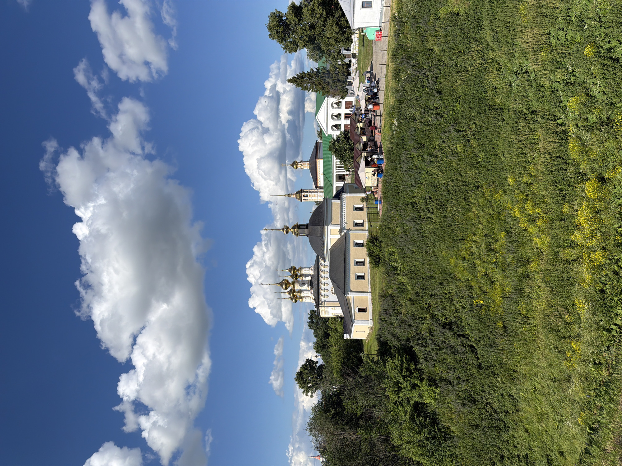 The image depicts a sunny day in Suzdal, Russia, with a view of the Suzdal Kremlin complex from a grassy hillside. The sky is a vibrant blue, dotted with fluffy white cumulus clouds. In the middle ground, several historic church buildings are visible, characterized by their golden domes and white, neoclassical architecture. Adjacent to the churches, there appears to be a small market or gathering area with stalls and umbrellas, where several people are congregated. The foreground is dominated by a lush, green hillside covered in dense vegetation and wildflowers, suggesting a natural or park-like setting. The overall scene conveys a sense of historical significance and a pleasant outdoor atmosphere.