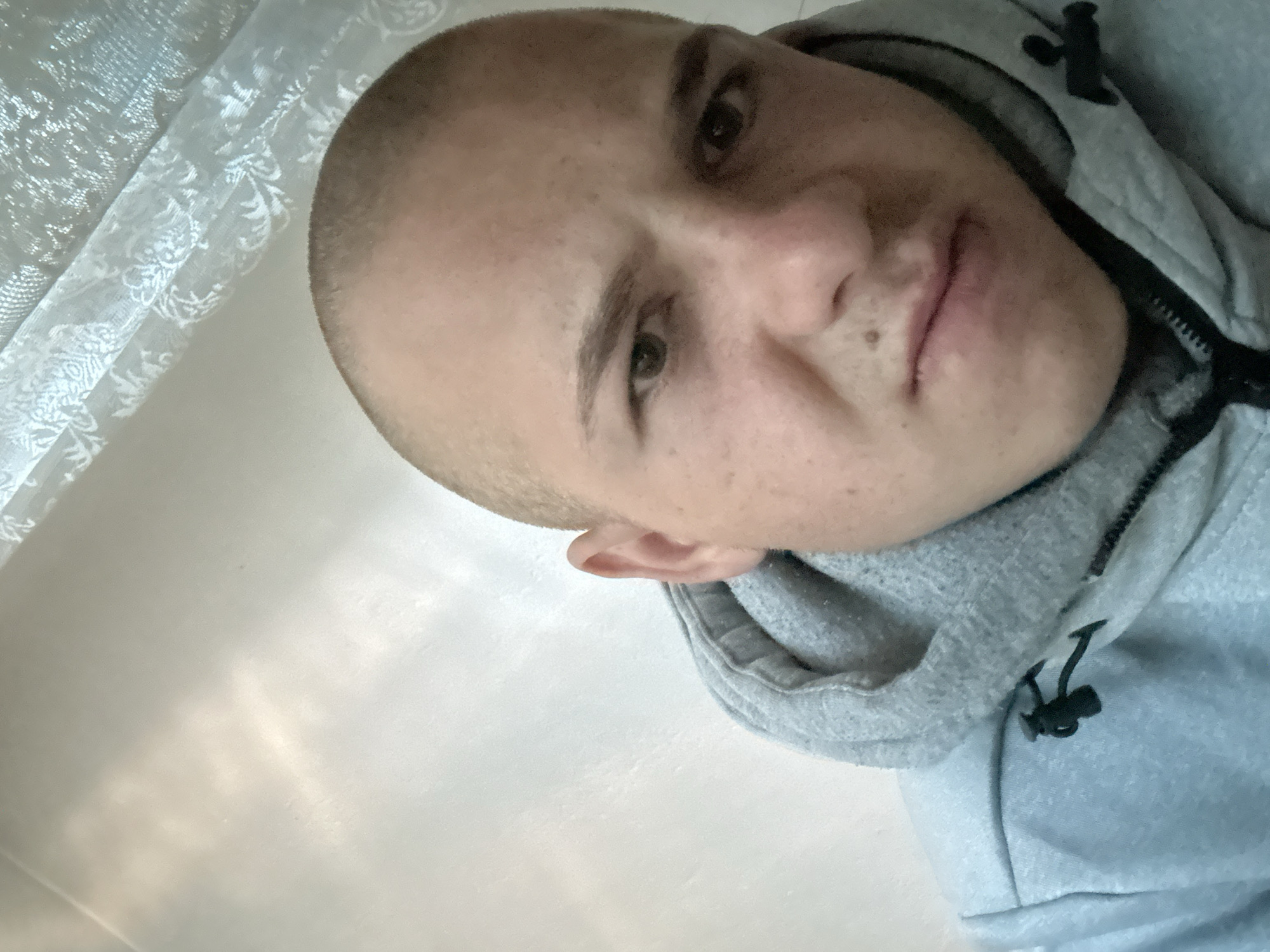 The image is a close-up, front-facing selfie of a person with a shaved head, wearing a light blue hooded sweatshirt. The person has light-colored skin, prominent eyebrows, and appears to be making a grimacing or disgusted expression. The background consists of a textured wall and a sheer white curtain with a patterned design. No specific location in Breznitsa, Bulgaria, can be identified from the visual information provided in the image.