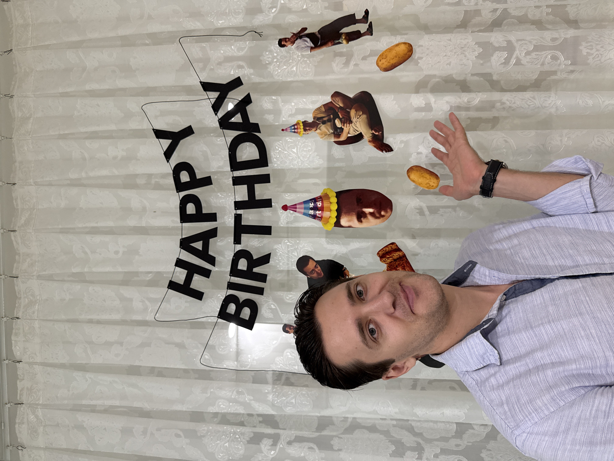 A medium shot shows a man in front of a white curtain. He wears a light blue button-up shirt and a black wristwatch. His arm is extended, with his palm facing the camera. Behind him is a transparent curtain with a printed "Happy Birthday" message. There are also cutout images of people and potatoes on the curtain, some wearing birthday hats. The location is suggested to be Nha Trang, Vietnam, but there is no visible information in the image to confirm this.