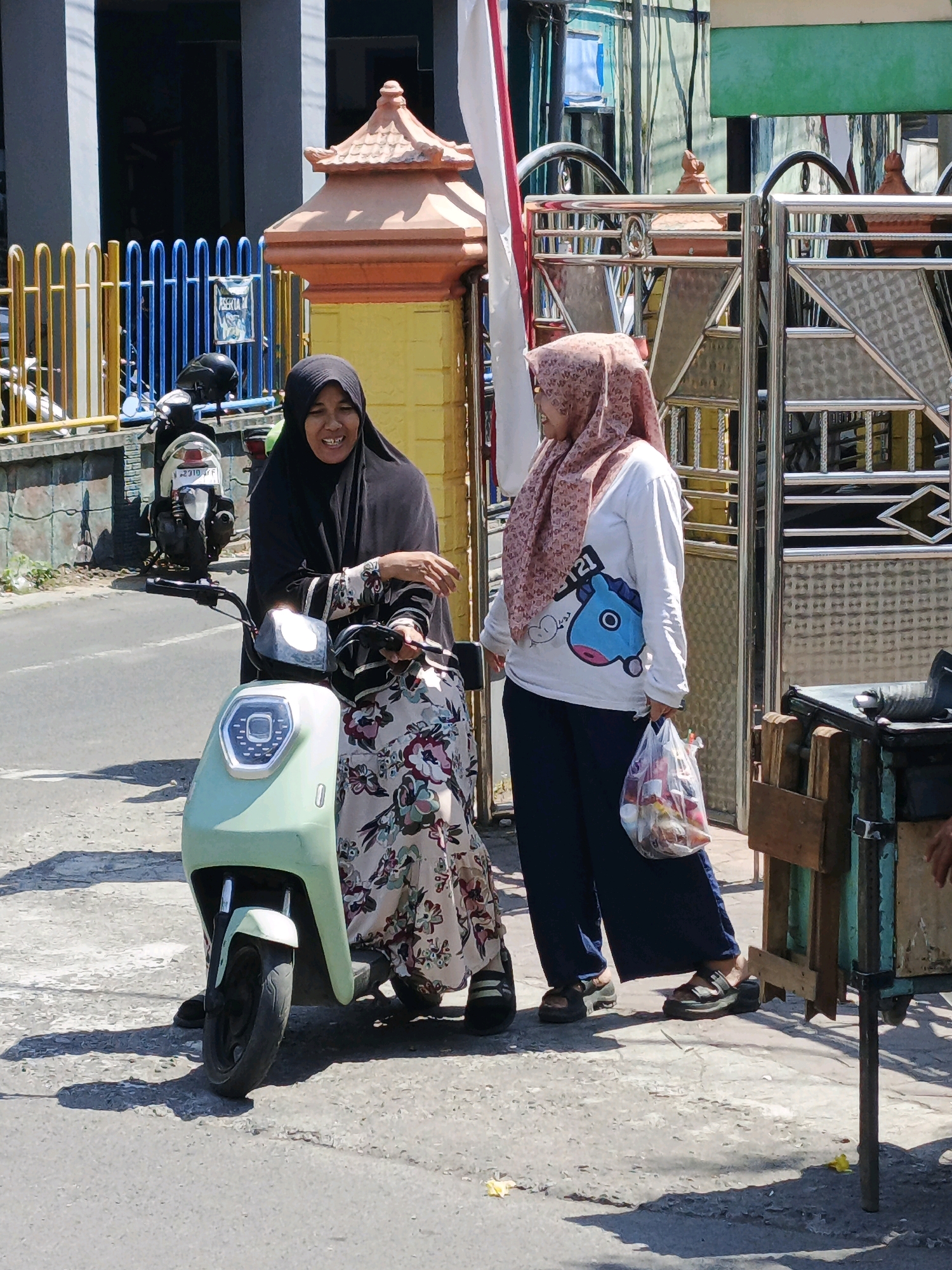 Two women stand on a sunlit sidewalk in front of a metal gate. One woman, wearing a black hijab and a patterned dress, sits on a light green electric scooter. She is smiling and looking towards the other woman. The second woman, wearing a patterned hijab and a white long-sleeved shirt with a blue cartoon character graphic, stands to the right of the scooter. She holds a plastic bag filled with various items in her right hand and is looking to her left. In the background, a blue fence and a yellow and orange decorative pillar are visible. The scooter has a license plate that reads "W 2310 TF". The scene appears to be in the city of Ketegan, Indonesia.
