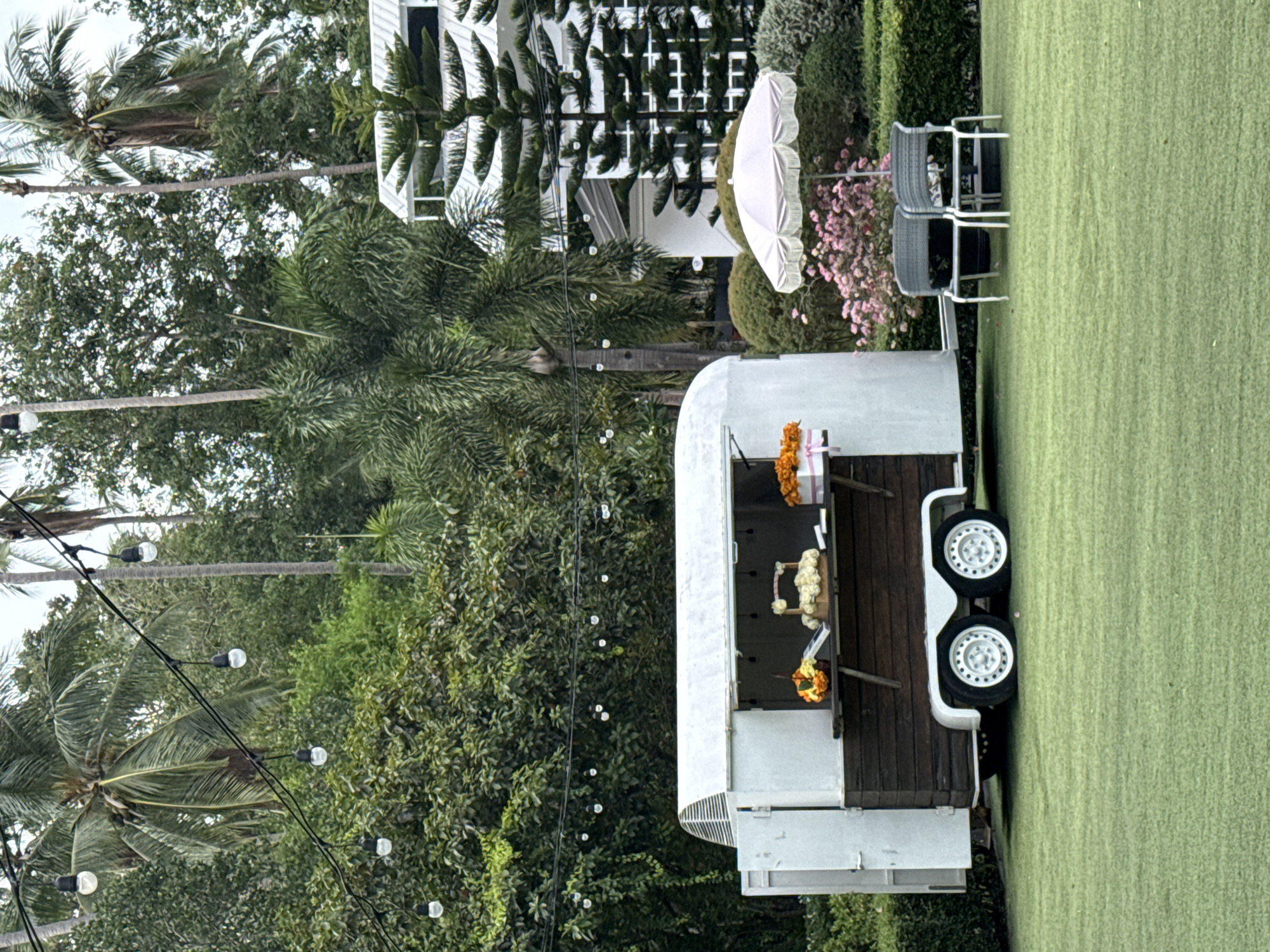 In the city of Ko Samui, Thailand, a food trailer sits on a meticulously manicured green lawn. The trailer, white with a wooden counter, is open, revealing a cake and floral decorations. To the right, a pale pink umbrella shades two chairs, with a backdrop of lush green trees and a white building. String lights are draped across the trees, suggesting an evening event.