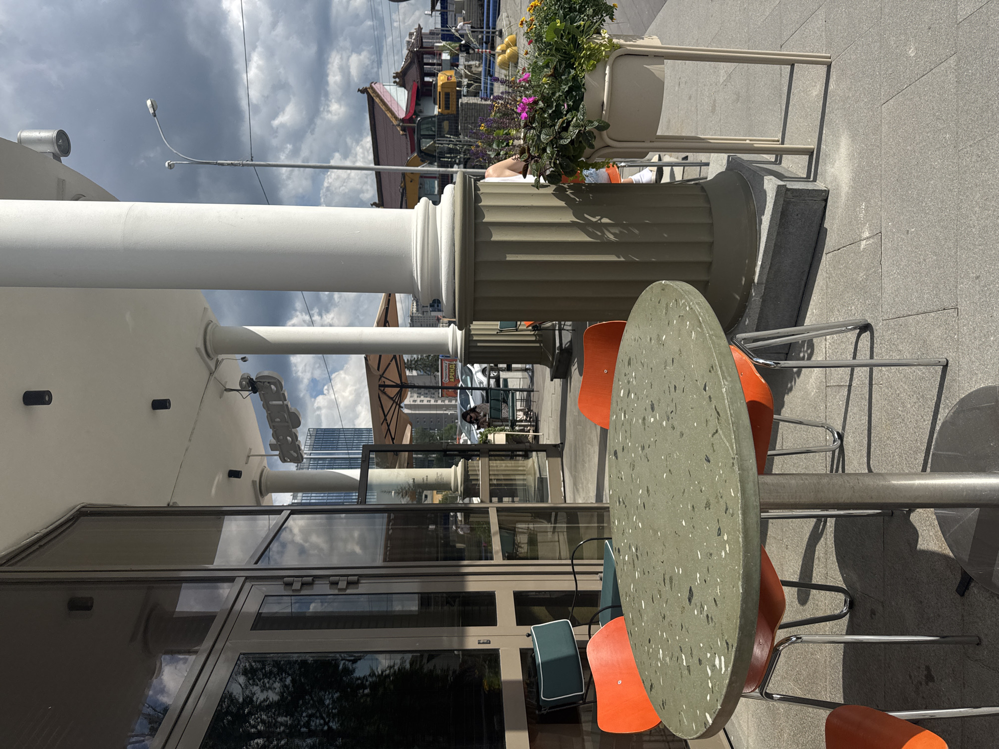 An outdoor seating area of a cafe is depicted on a cloudy day in Yekaterinburg, Russia. In the foreground, a round, speckled green table with a silver base is surrounded by four bright orange chairs with silver legs. To the right of the table, a tall, cream-colored planter holds an arrangement of purple and yellow flowers. Behind the planter, part of a white column is visible. The cafe's facade features large glass windows and doors, reflecting the sky and surrounding buildings. Further back, additional outdoor seating with tables and chairs can be seen, along with an open umbrella. In the background, a street scene unfolds with parked cars, construction equipment including a yellow excavator, and various buildings, one of which appears to be a traditional Asian-style structure with a red roof. The sky is filled with dramatic, dark clouds.