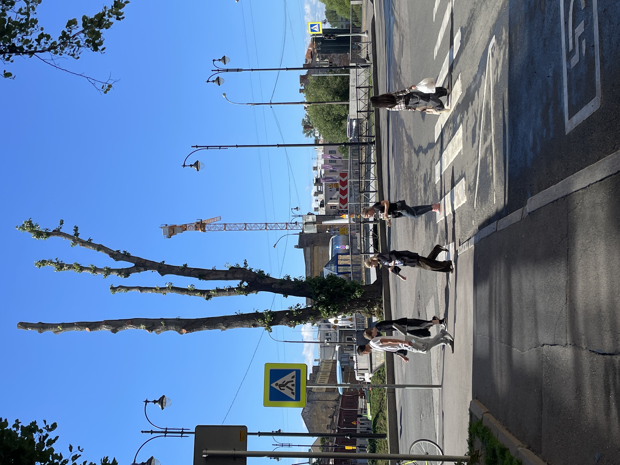 A group of five young adults are crossing a pedestrian crosswalk on a sunny day in Saint Petersburg, Russia. The scene is captured from a slightly elevated perspective, looking down at the crosswalk and the surrounding urban environment. The sky is a clear, bright blue, suggesting fair weather.
The individuals are dressed casually, and their body language indicates they are walking across the street. One person in the front is wearing a black top and light-colored pants, with their back to the camera. Behind them, another individual is wearing a white t-shirt and grey pants. Further back, a person in a dark outfit is walking alongside another in a plaid shirt and dark pants, carrying a white bag. The last person visible is in a dark outfit, walking slightly ahead.
The street is paved with asphalt, and the crosswalk is marked with white stripes. Shadows are cast across the road, indicating the sun is high, likely midday. Streetlights with multiple lamps are positioned along the sidewalk. In the background, there are buildings with a mix of architectural styles, some appearing older and some more modern. A construction crane is visible in the distance, suggesting ongoing development or renovation in the area. A yellow pedestrian crossing sign with a blue border and a silhouette of a walking person is prominently displayed on the left side of the image. The overall impression is a typical urban scene on a pleasant day, with people going about their daily activities.