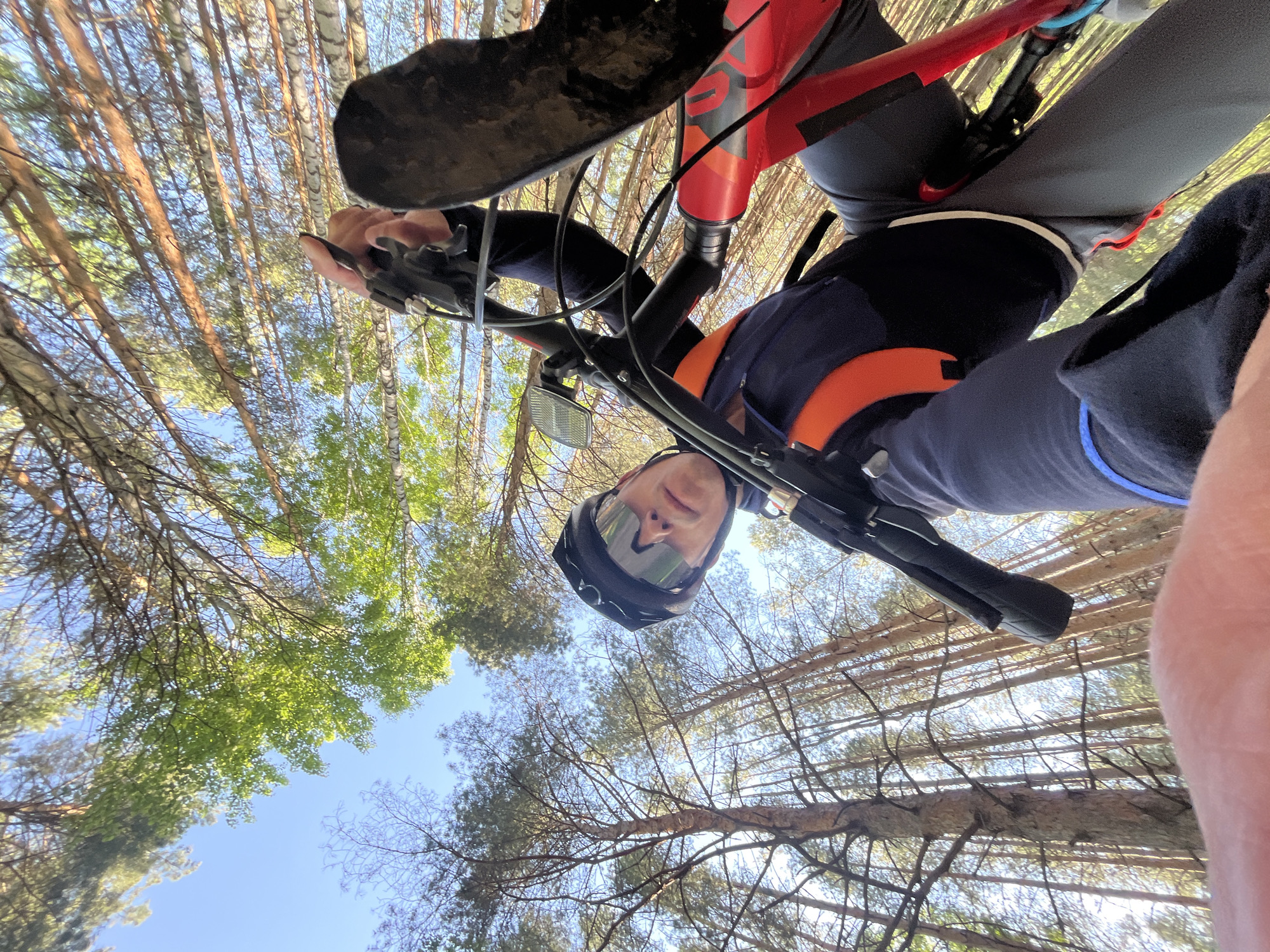 A cyclist wearing a helmet and sunglasses, possibly a selfie, is captured from a low-angle perspective looking upwards. A red and black mountain bike is partially visible, with the handlebars and front tire occupying the upper portion of the frame. The cyclist is wearing a dark blue top with an orange strap and a blue stripe on their sleeve. The background consists of a forest of tall trees, primarily pines and birches, with a blue sky visible through the canopy. This photo was taken in a forest in the city of Chaikovsky, Russia.