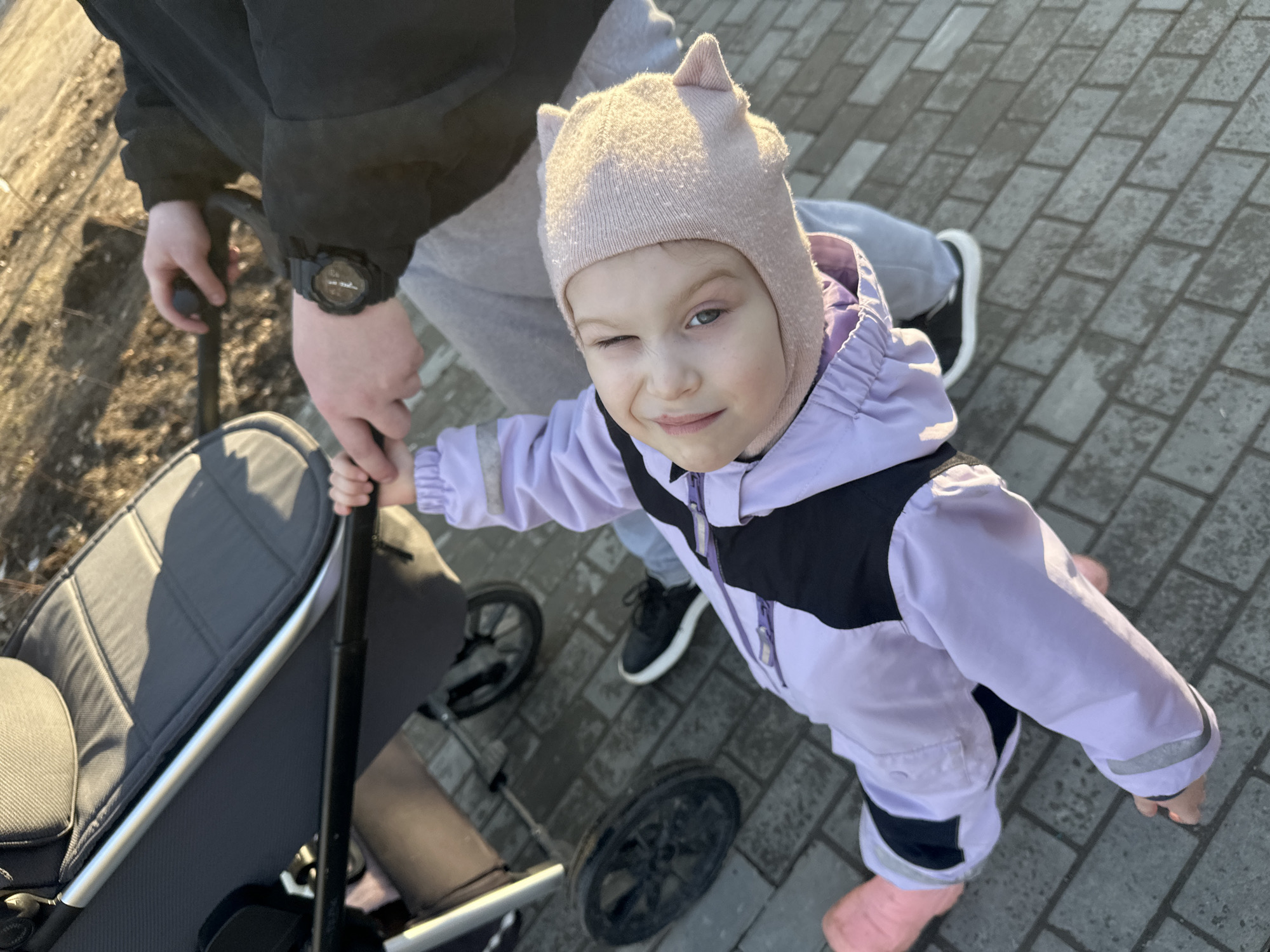 A young child winks at the camera while standing on a paved surface near a stroller being pushed by an adult. The child is wearing a light purple jacket, a pink hat with pointed ears, and pink boots. The adult is wearing a dark jacket, gray pants, and black and white sneakers. The location appears to be an outdoor area within the city of Chaikovsky, Russia. The stroller is dark gray with a silver frame and a black handle. The paved surface is made of gray bricks. The photo seems to be taken during the day with natural lighting.