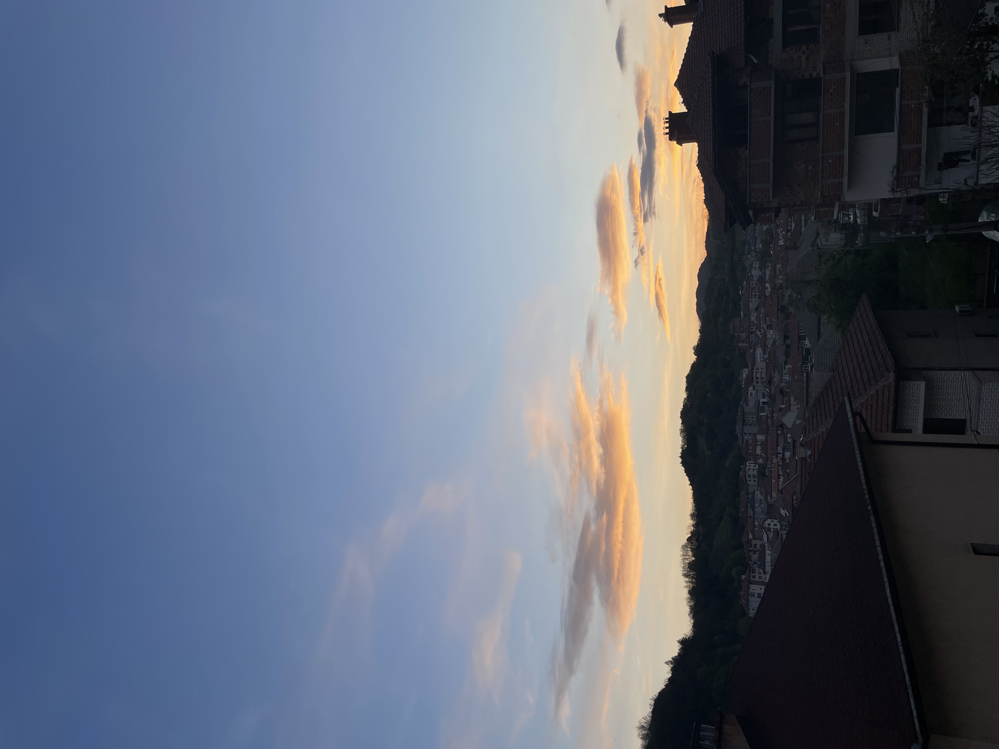 The image presents a view of Zlatograd, Bulgaria, captured from an elevated position, possibly a rooftop. The lower portion of the frame includes parts of buildings, with a reddish-brown tiled roof being prominently visible in the foreground on the right side of the image. A building with exposed brickwork and window openings is visible in the upper right corner of the image, indicating construction or renovation.
A forested hill line borders the town. Between the foreground elements and the hillside is an urban area consisting of low-rise buildings with red tile roofs. The upper left area of the image is dominated by a blue sky with elongated, orange-tinted clouds that are concentrated closer to the horizon. The lighting suggests that the image was taken during sunset or sunrise.