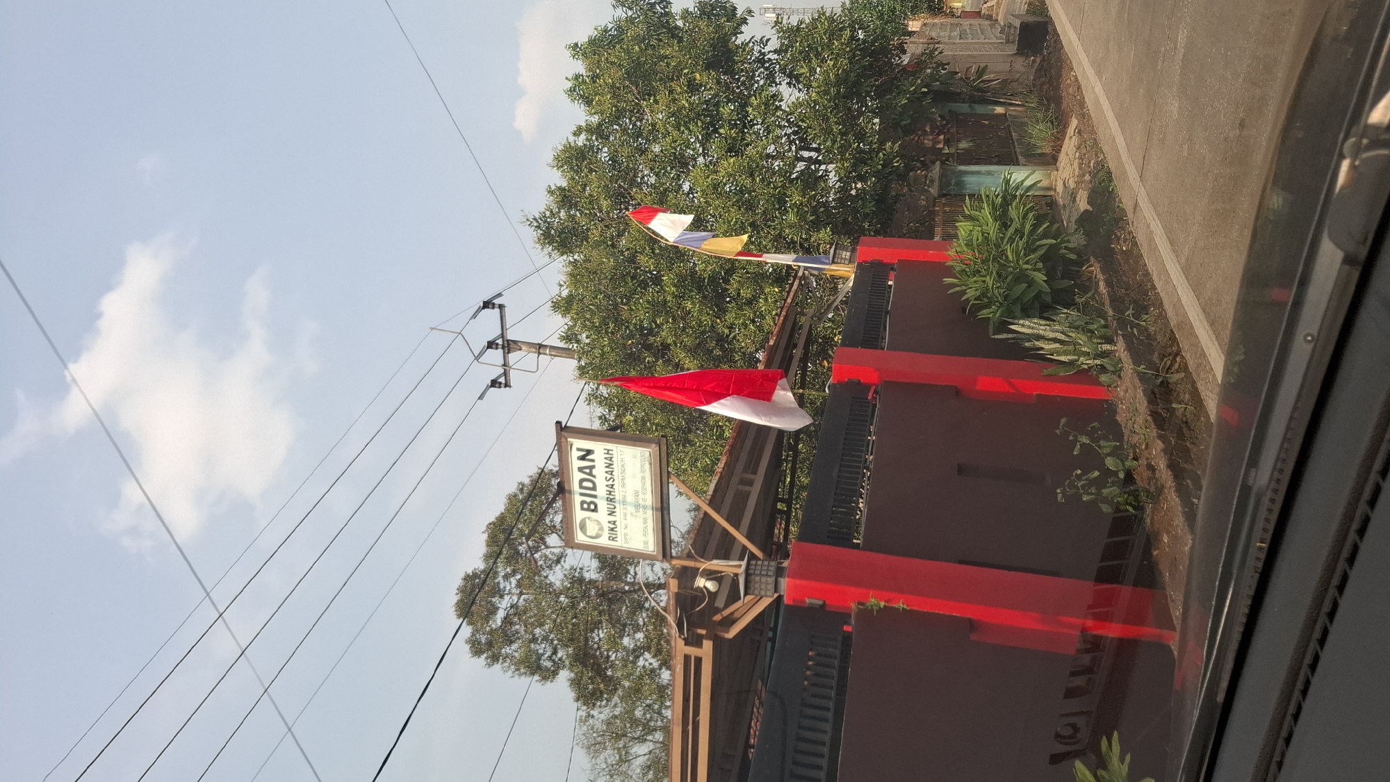 The image shows a street scene in Cibinong Hilir, Indonesia. A red and white Indonesian flag is flying on a pole in front of a building with a sign that reads "BIDAN RIKA NURHASANAH". The building is red and has a black fence. There are trees in the background and power lines overhead. The image appears to have been taken from a car.