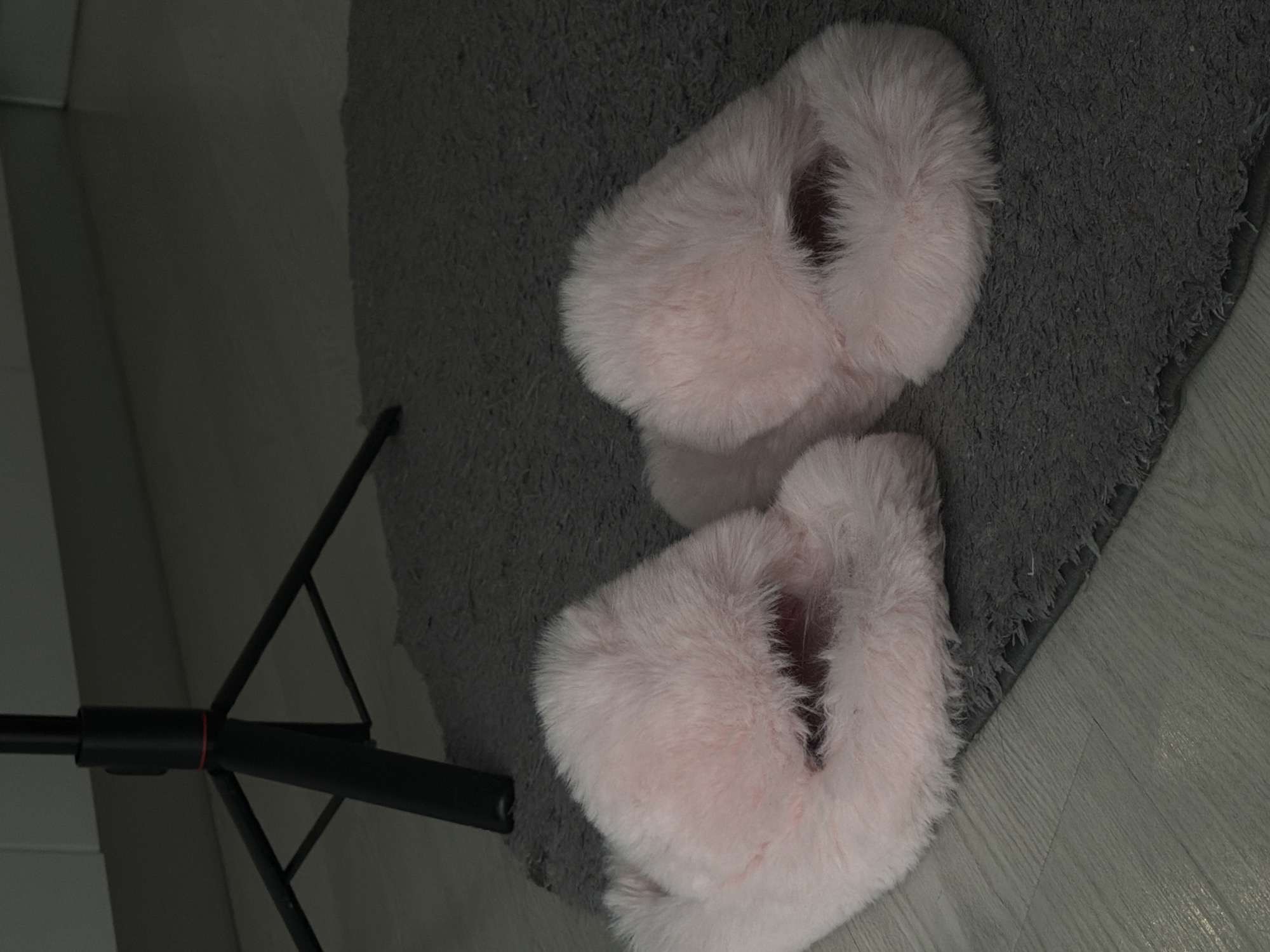 This image captures a rotated overhead view of an indoor domestic scene in Sofiivska Borshchahivka, Ukraine. The main subjects are two light pink, fluffy slippers resting on a dark gray shaggy rug. The slippers appear soft and plush, suggesting comfort and warmth.
The rug has a thick, textured pile and is laid on a light-colored floor, which appears to be wood laminate or tile with a visible grain pattern. A black metal tripod stand is partially visible on the left side of the frame, with its slender legs and central joint extending over both the floor and the rug. A light-colored wall or piece of furniture is also visible along the far left edge.
No activity is taking place; the slippers are simply positioned on the rug. The lighting is soft and ambient, characteristic of an indoor setting. The scene evokes a sense of comfort and relaxation within a private home environment. There is no visible text in the image.